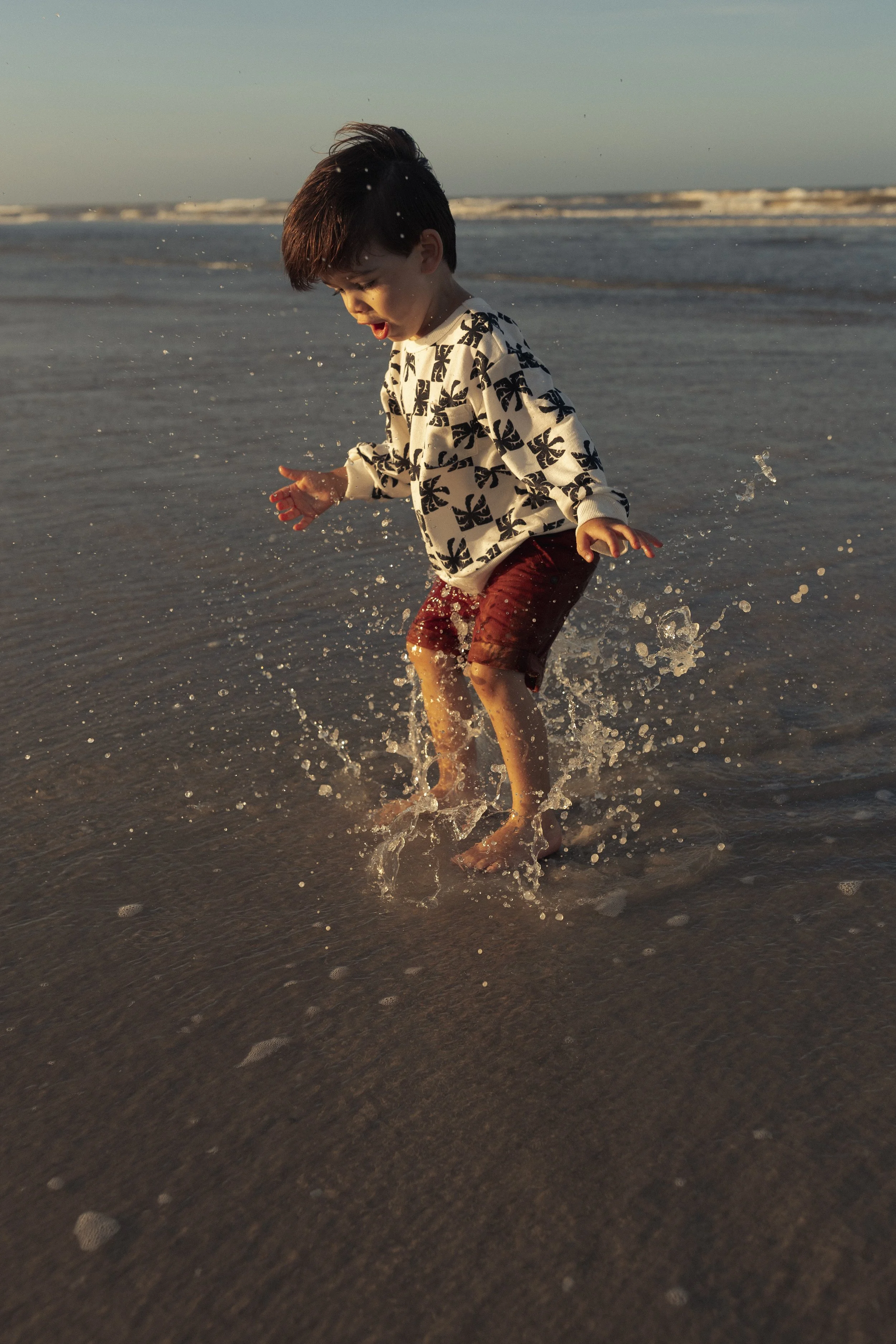 A young boy playing and splashing in the shallow ocean water on the beach during sunset.