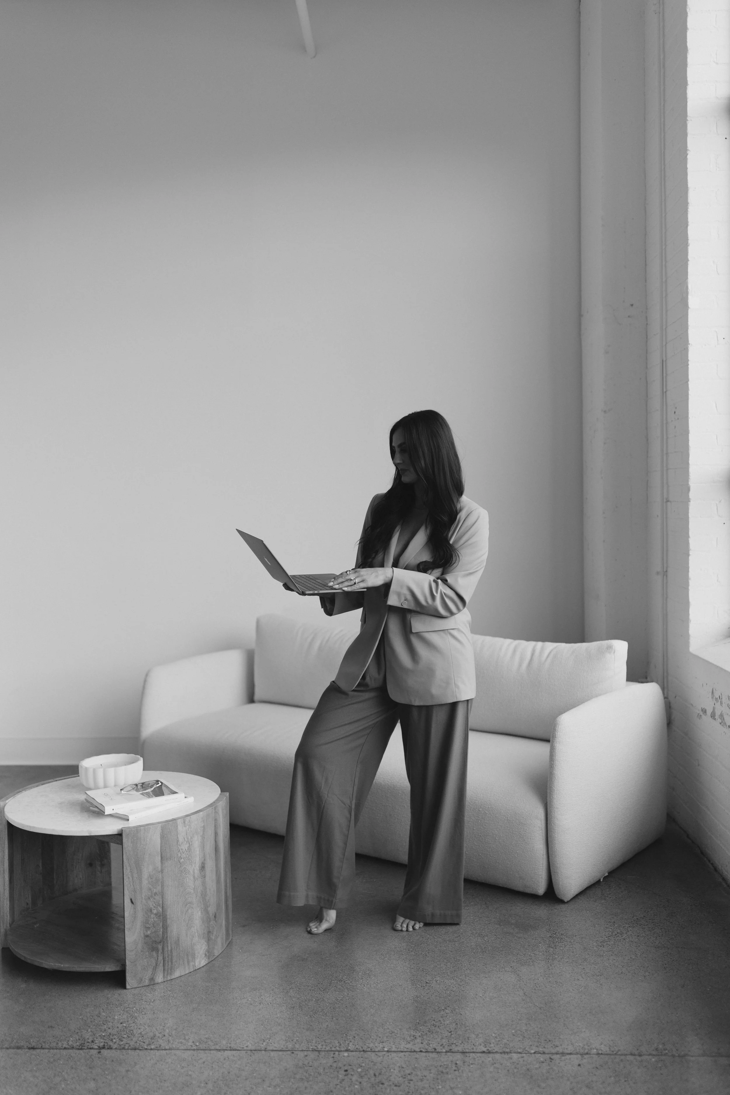 A woman in a blazer and wide-leg pants standing barefoot in a minimalist living room, looking at her laptop, with a sofa and a small round wooden table nearby.