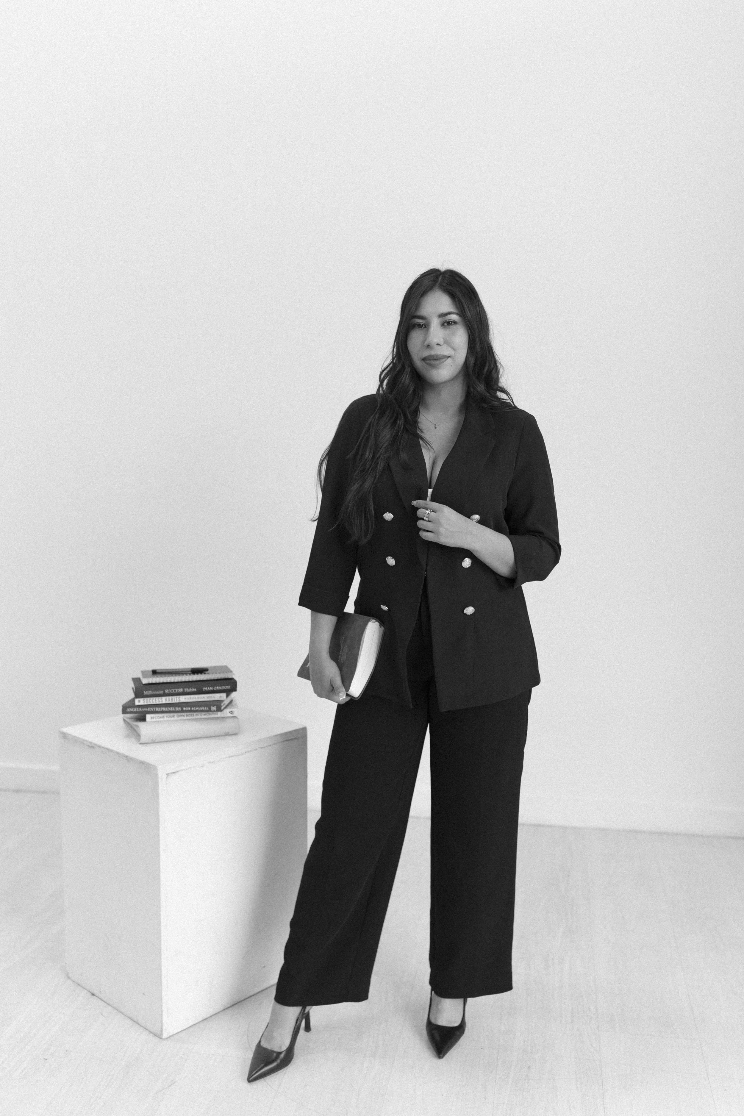 A woman in a black suit with wide-leg pants and pointed heels standing next to a small white cube with a stack of books and a phone on top, holding a book and looking at the camera.