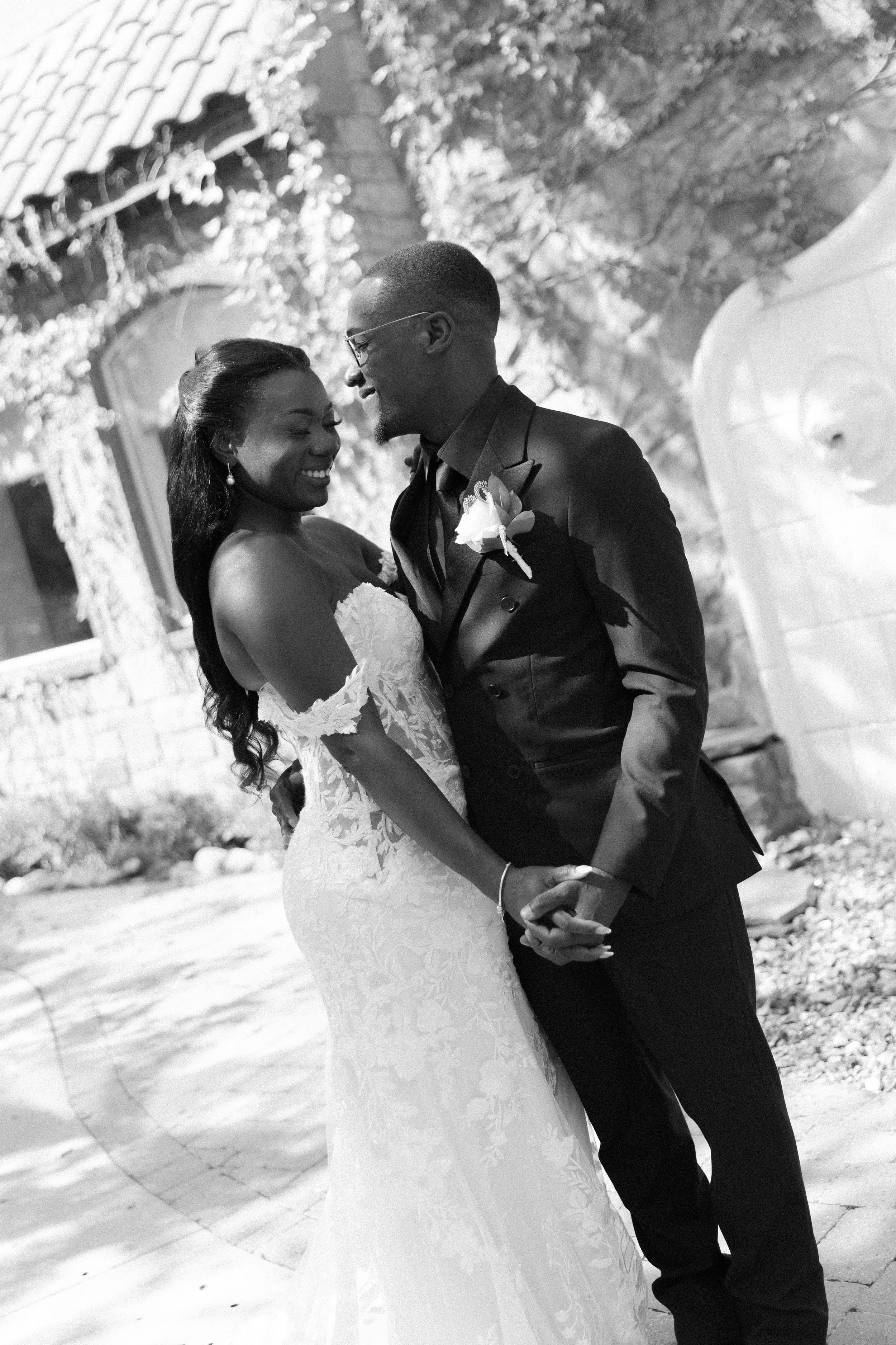 A black and white photo of a bride and groom smiling and holding hands during their wedding. They are standing close to each other outdoors, with a stone building and landscaping in the background.