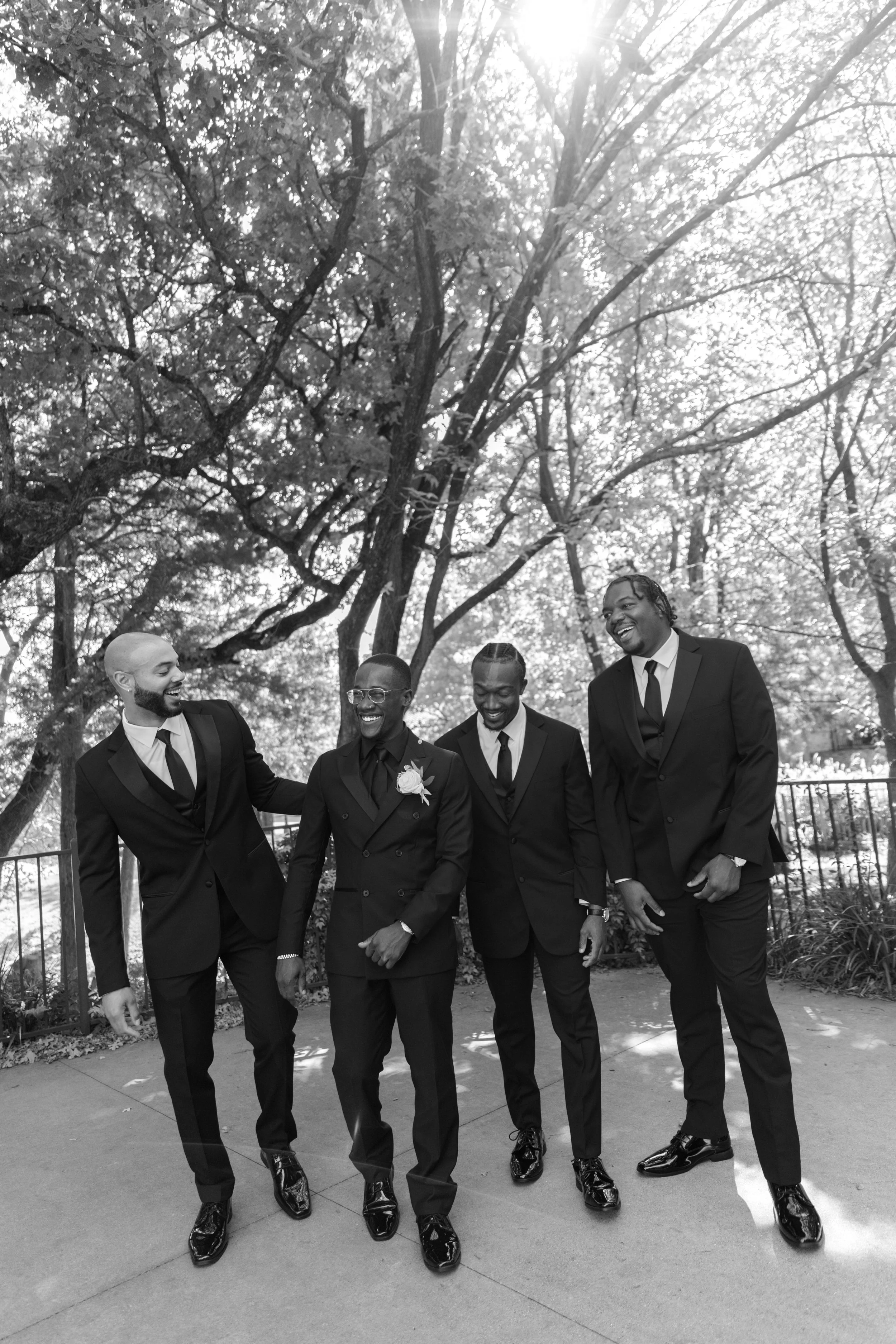 Four men dressed in formal suits walking outdoors, laughing, under a large tree with sunlight filtering through the leaves.