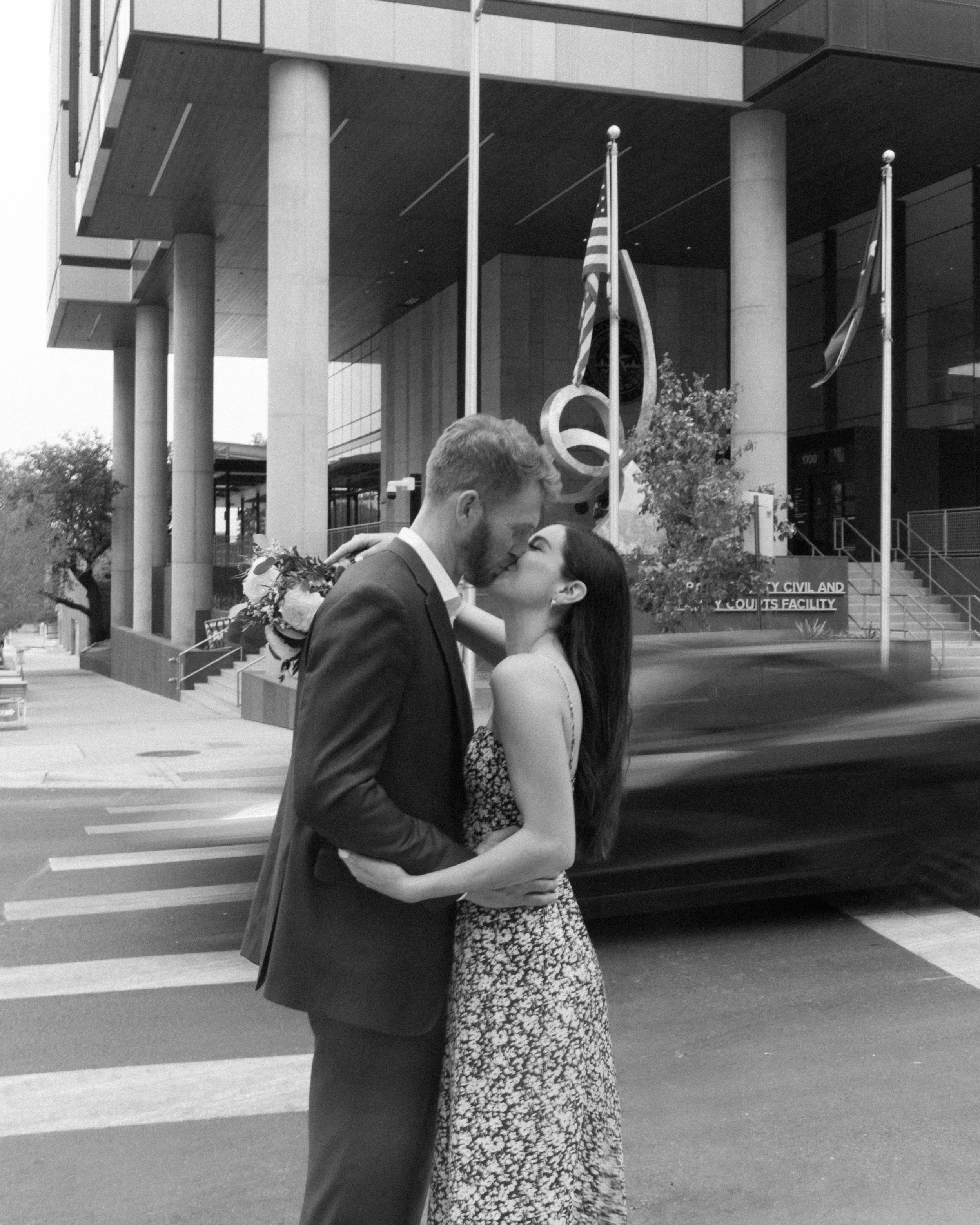 A black and white photo of a couple kissing in the street in front of a modern building, with flags and trees in the background.