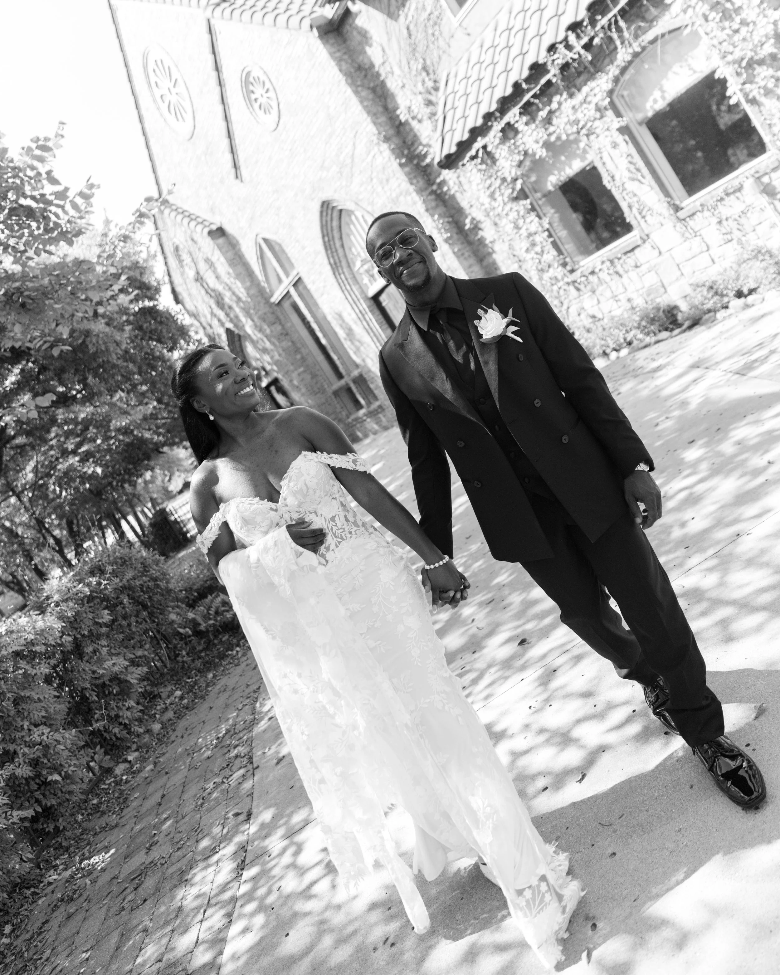 A black-and-white photo of a smiling bride and groom holding hands outdoors in front of a historic brick building with arched windows, trees, and a cobblestone path.