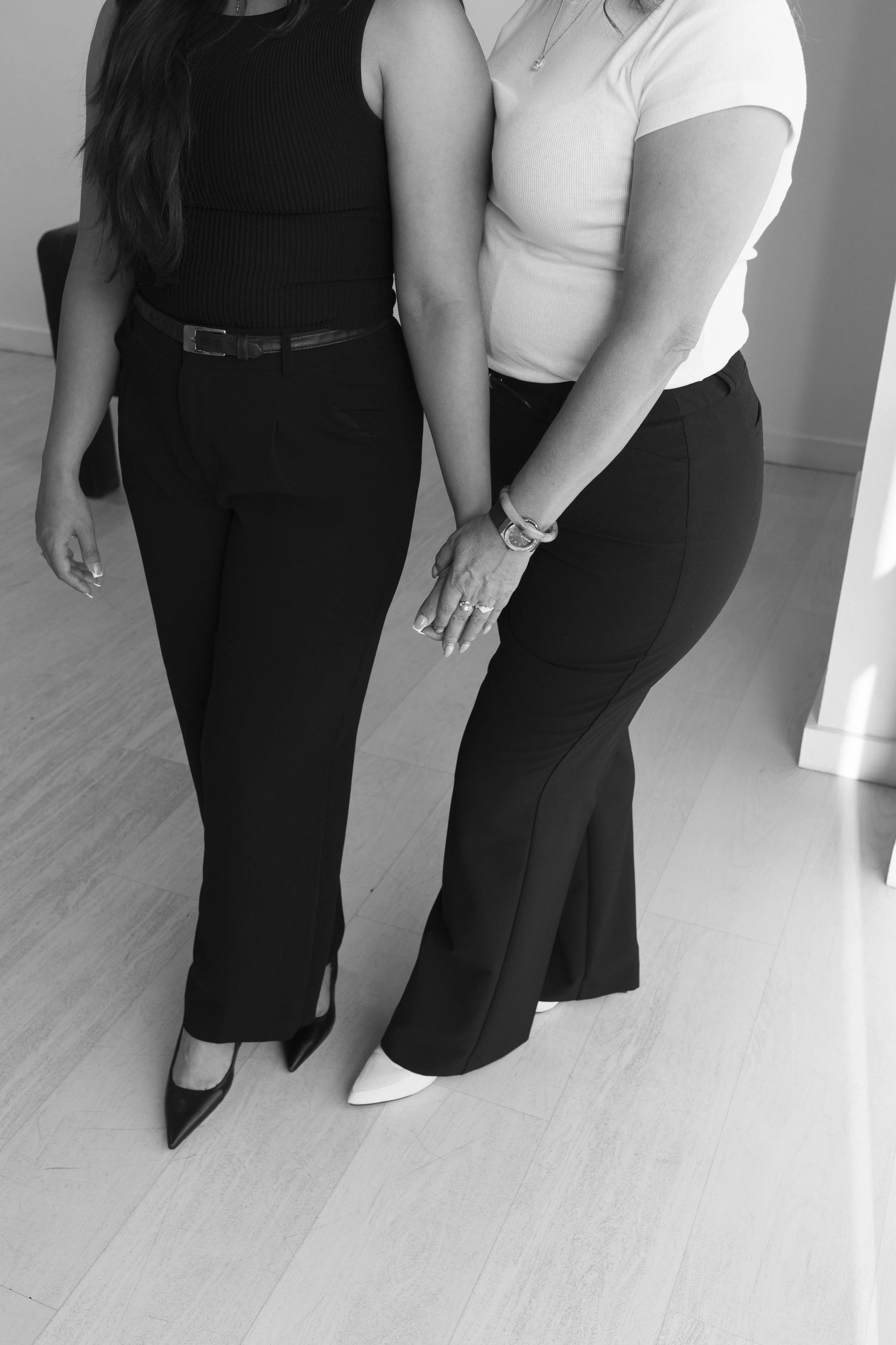 Black and white photo of two women standing close together, holding hands with fingers interlocked, wearing high heels and fitted pants and tops.