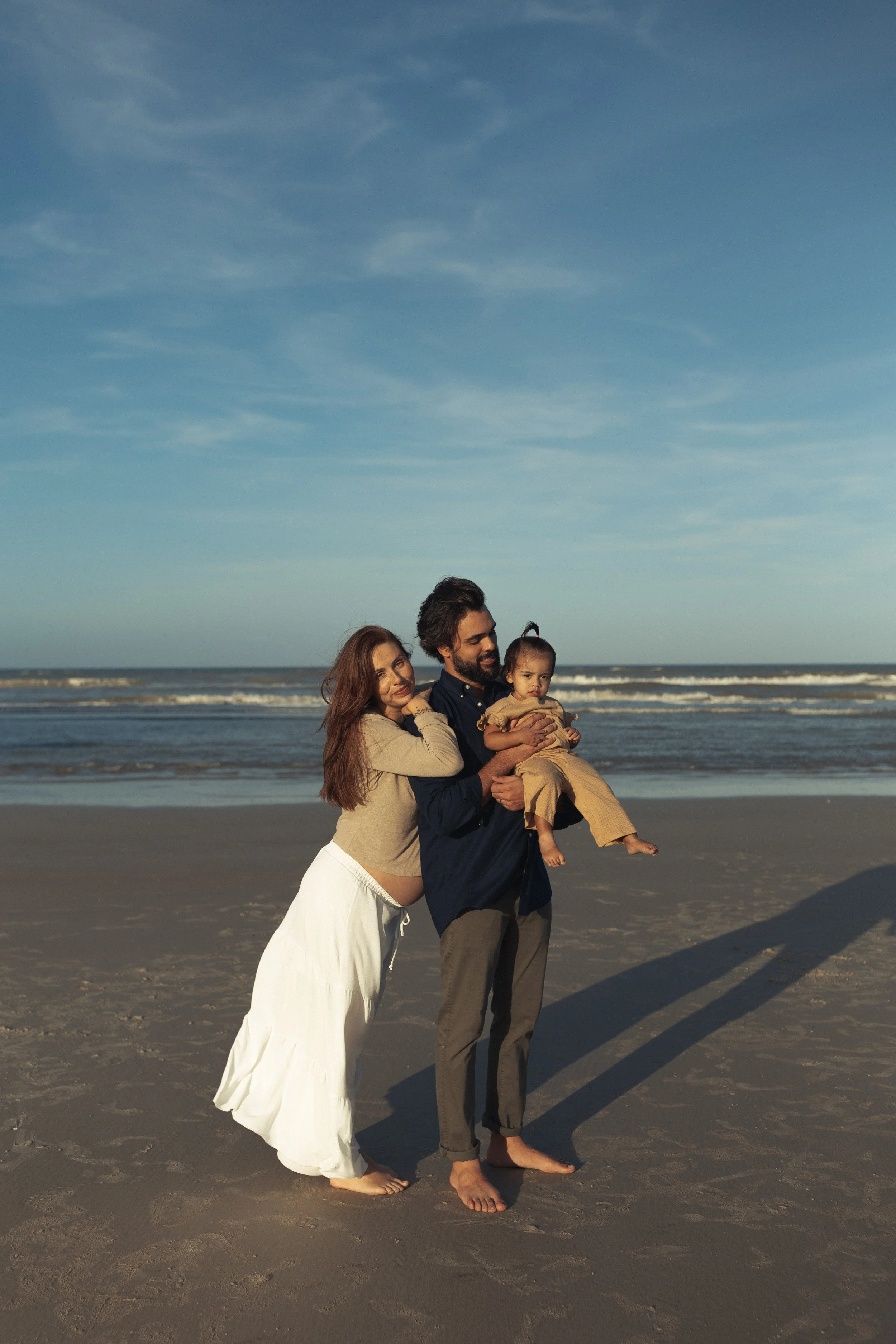 Family of three enjoying time at the beach, with the father holding a young girl and the mother leaning on him, all standing barefoot on the sand under a blue sky.