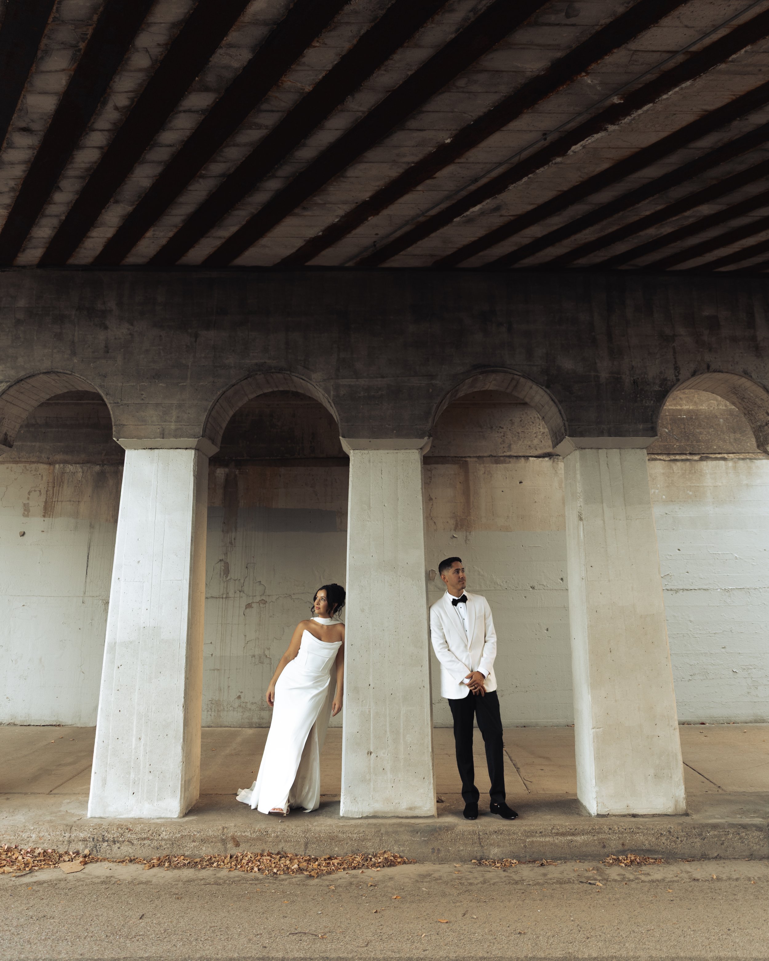 A woman in a strapless white gown and a man in a tuxedo stand in an urban, concrete underpass, separated by concrete columns.