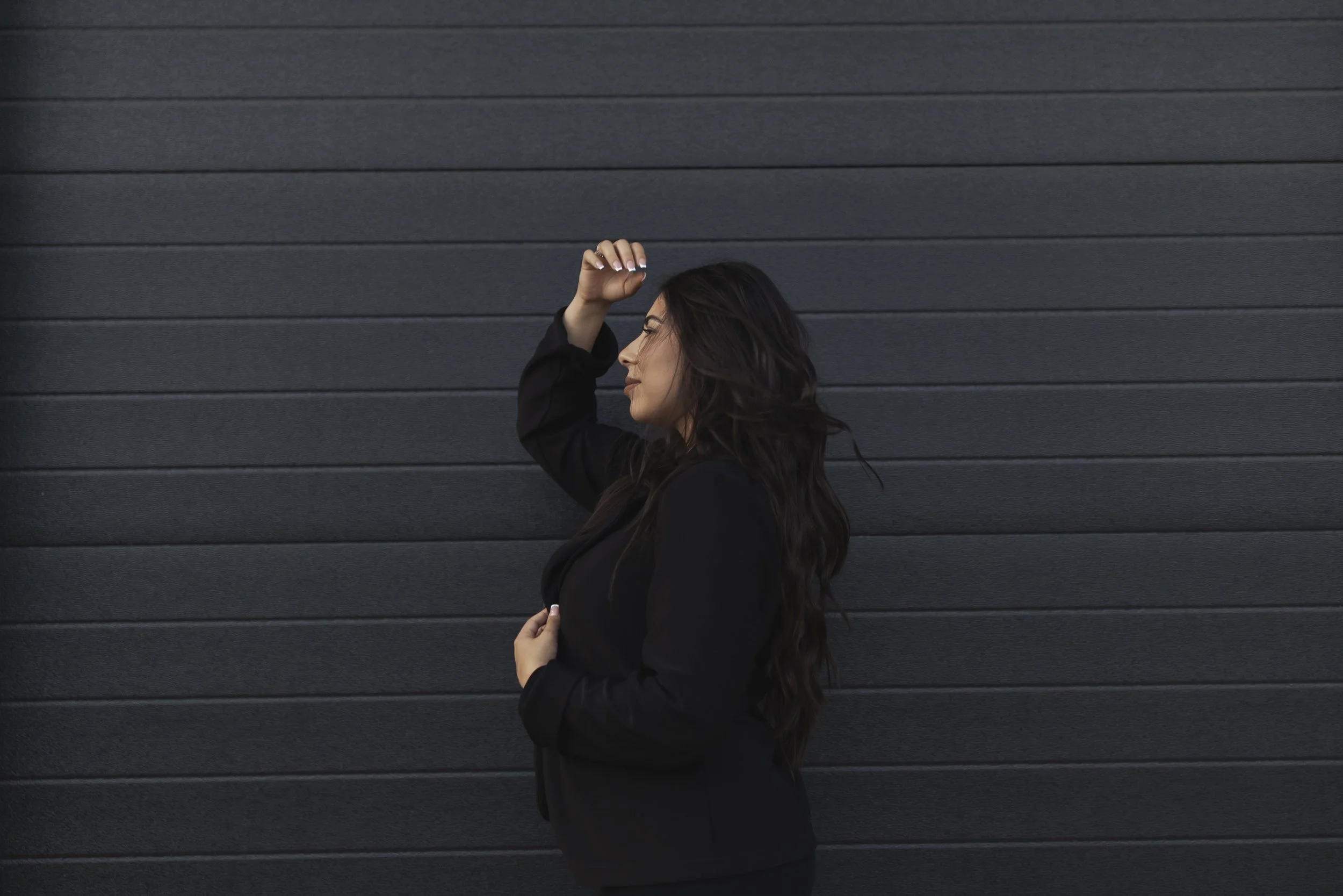 A woman with long dark hair dressed in black standing sideways in front of a black corrugated metal wall, with her eyes closed, using one hand to hold her blazer and the other raised to her forehead.