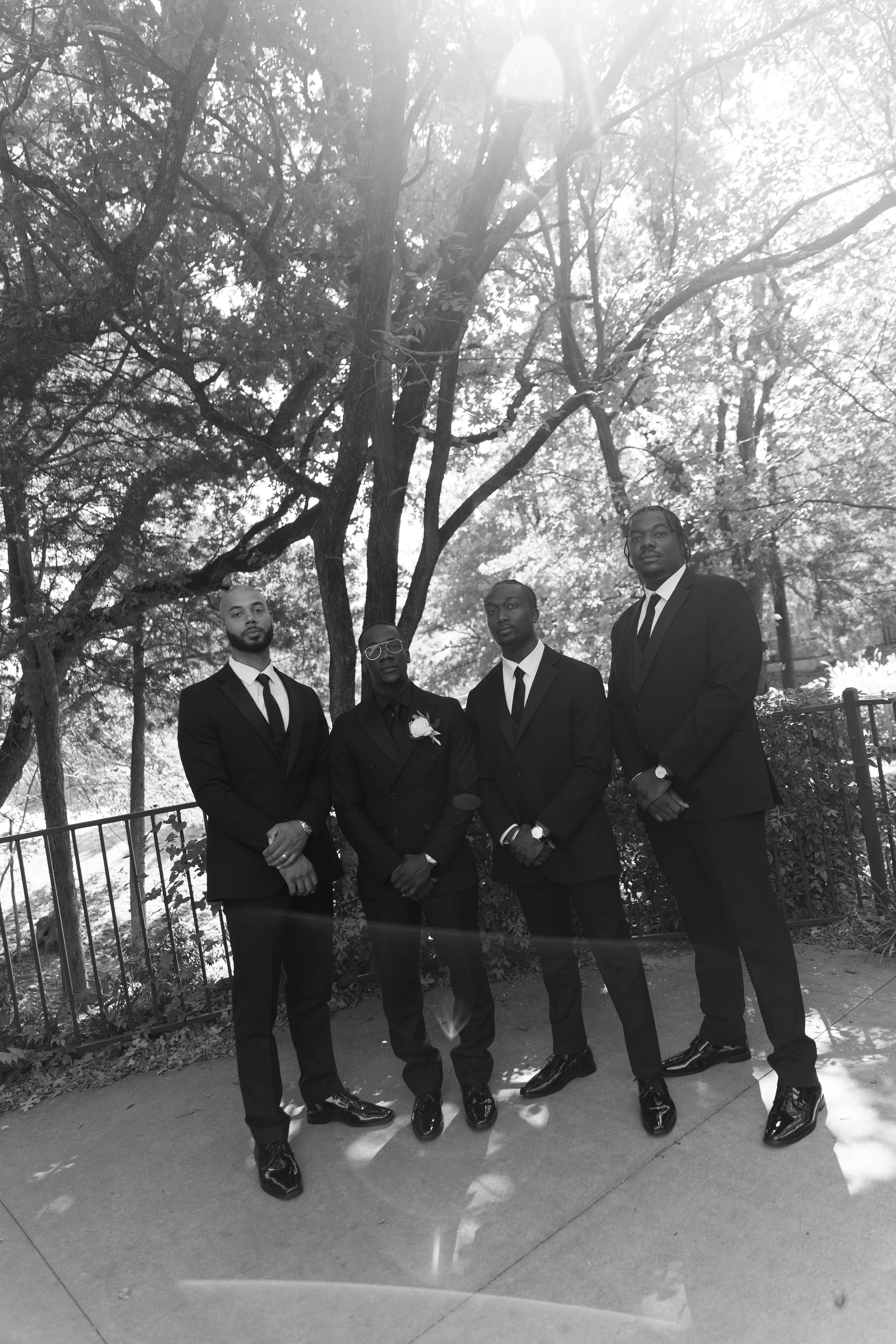 Four men dressed in formal black suits with ties, standing outdoors on a sidewalk near trees, with bright sunlight filtering through the branches, one wearing glasses and a flower boutonniere.