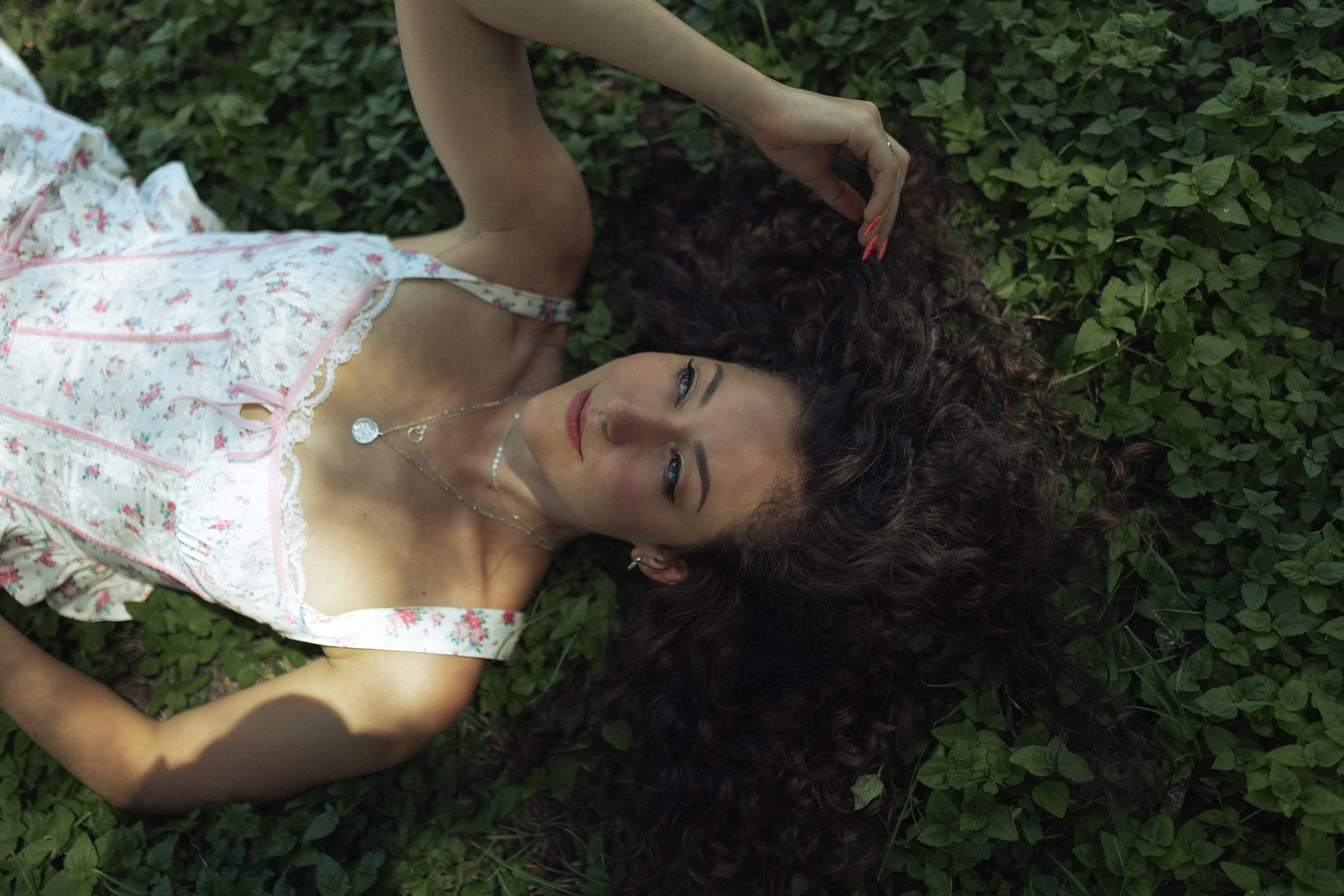 A woman with curly hair is lying on a bed of green leaves, wearing a floral nightgown and looking up.