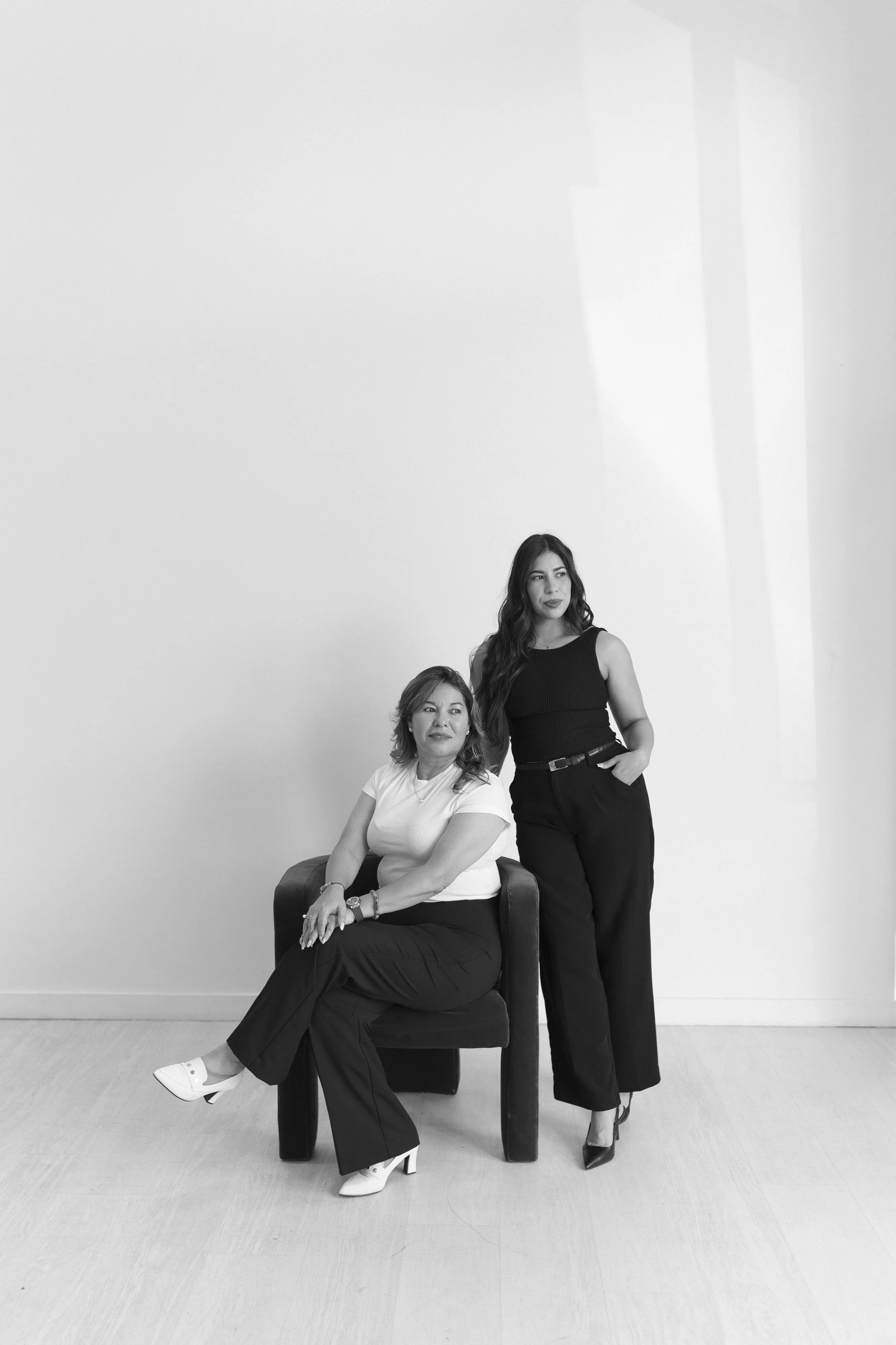 Two women in black and white, one sitting on a chair and the other standing beside her, both looking off to the side in a minimalistic room.