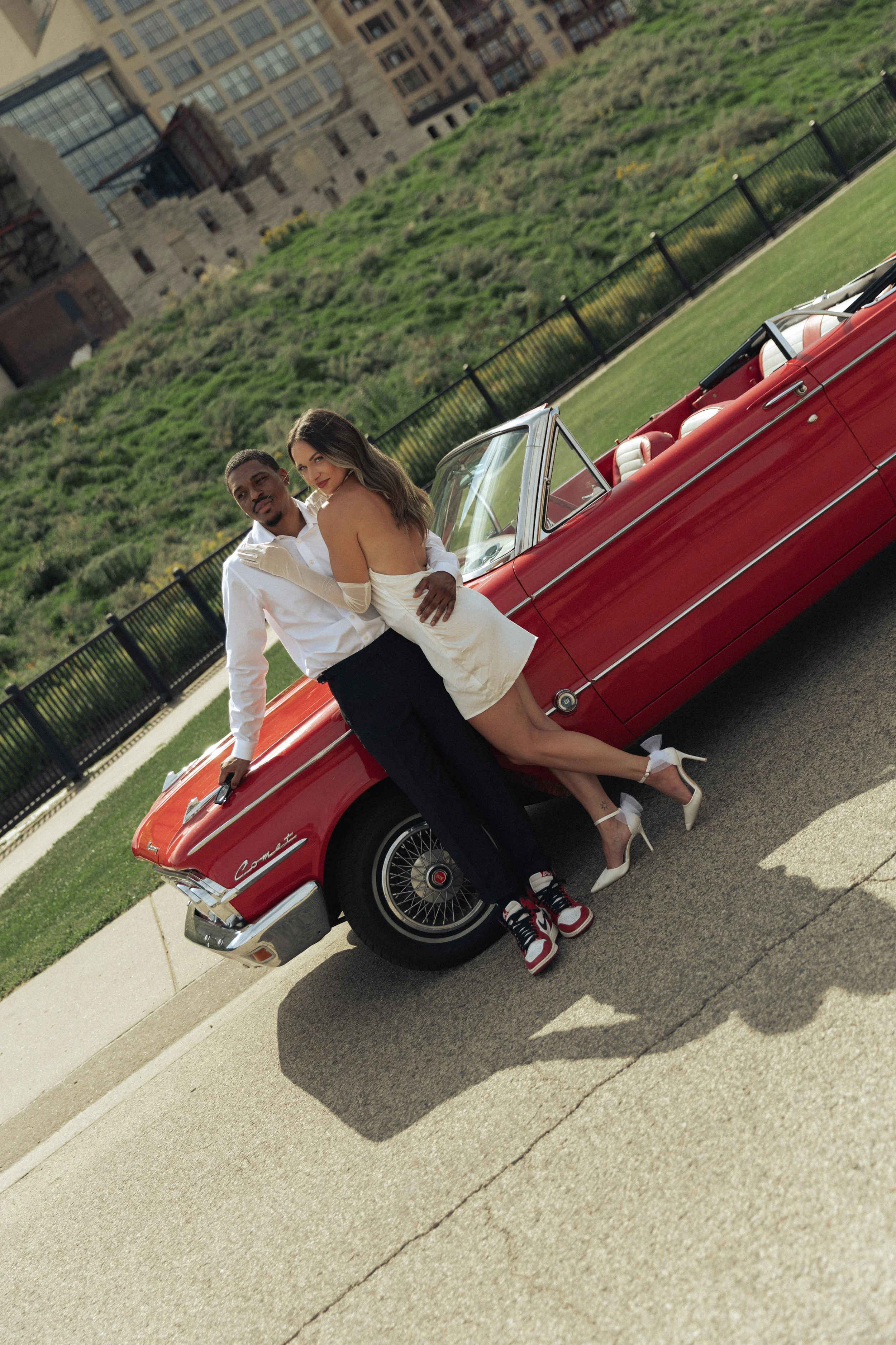 A man and woman in stylish attire leaning against a red vintage convertible car outdoors in a park with grass, fencing, and city buildings in the background.