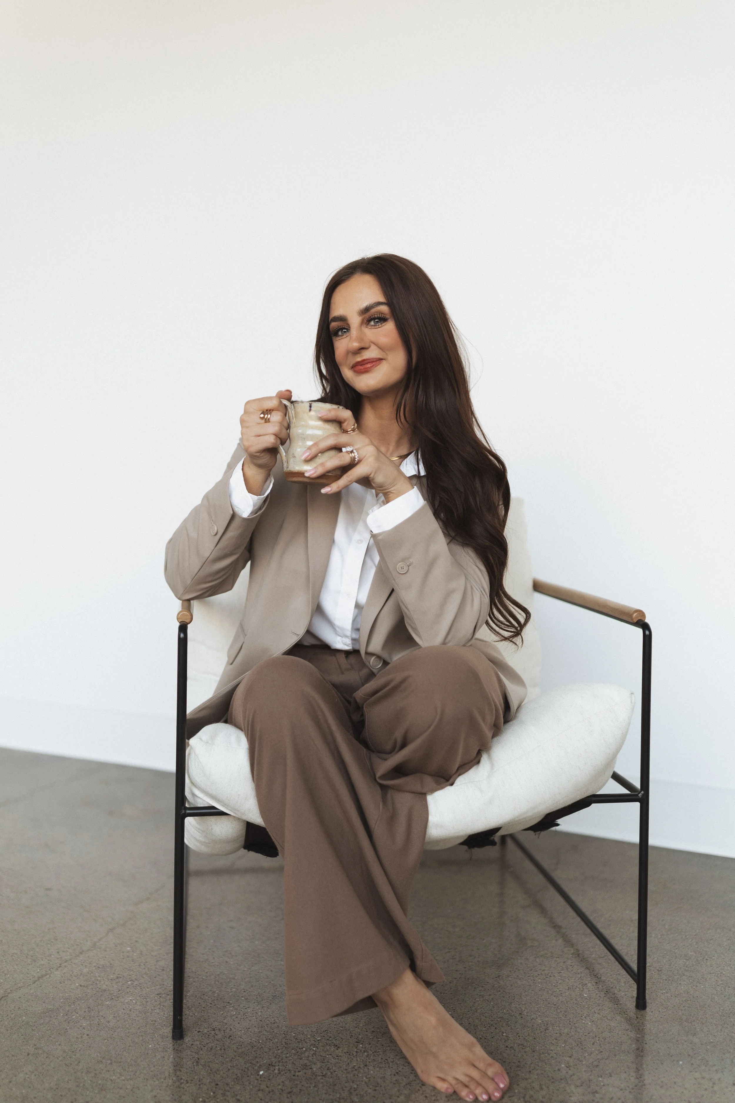 A woman with long brown hair sitting on a modern cushioned chair, holding a ceramic mug, wearing a beige blazer, white shirt, and brown wide-leg pants, against a plain white background