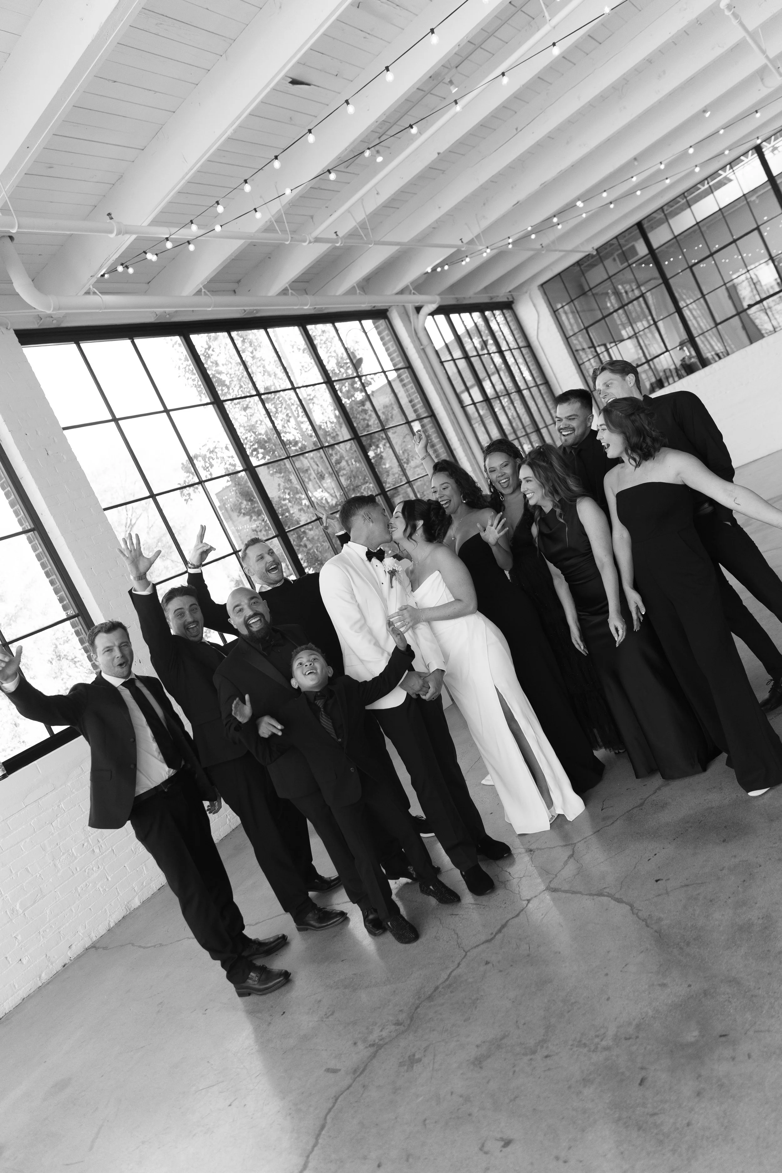Black and white photo of a large group of people in formal attire, celebrating indoors with a high, ceiling, string lights, large windows, and a brick wall.