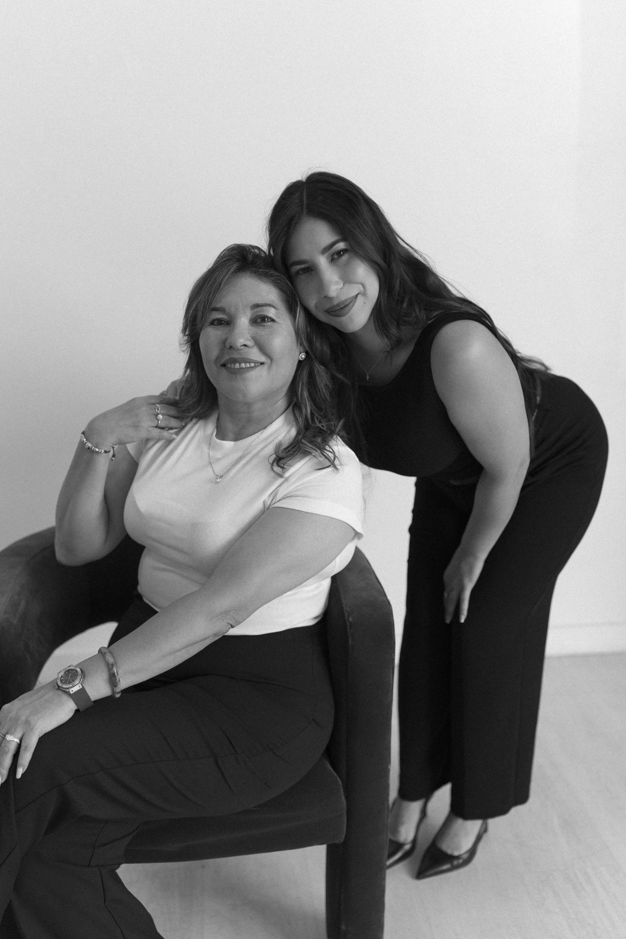 Two women, one seated in an armchair and the other standing beside her, smiling at the camera in a black and white photo.