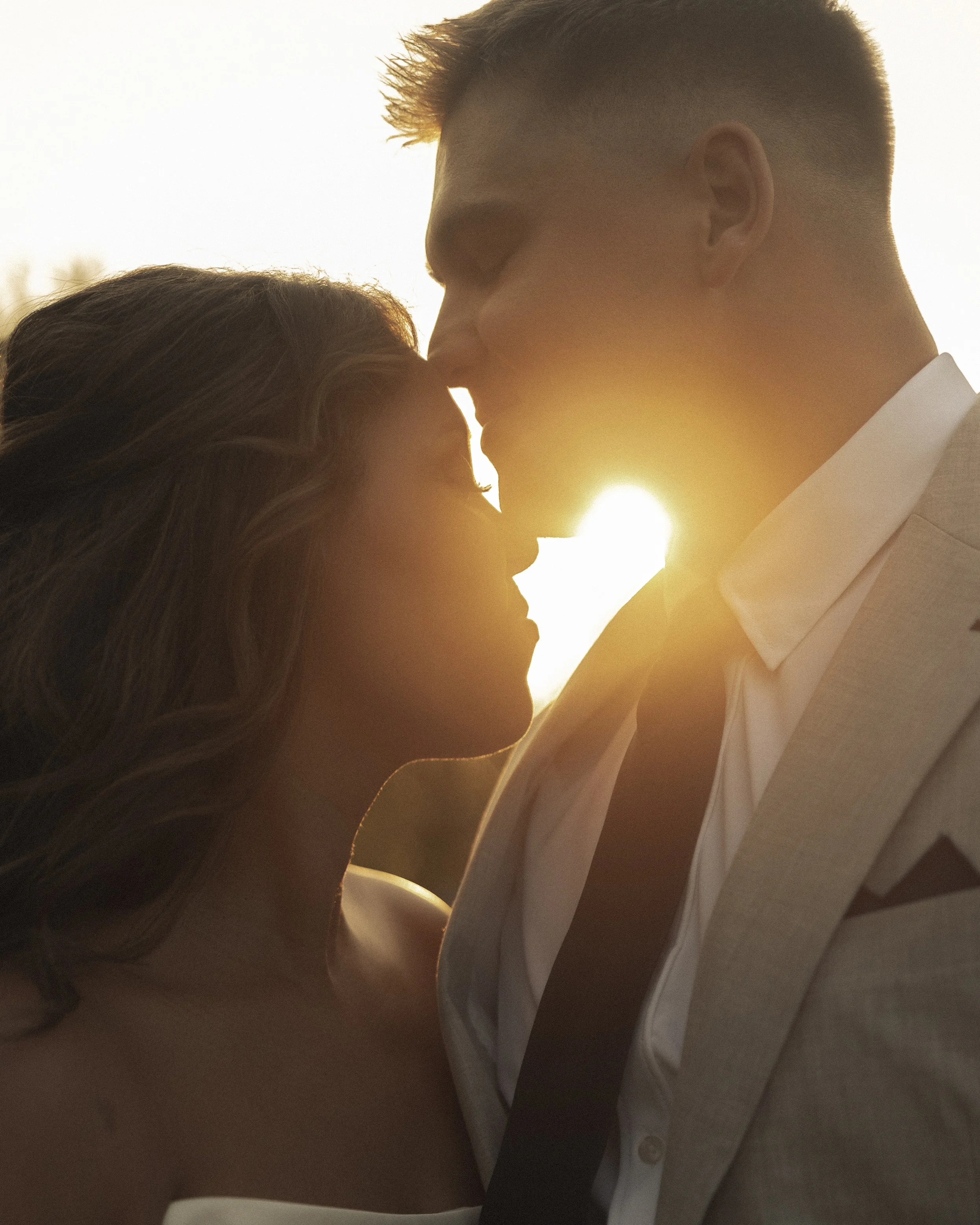 A couple is standing close together, with their foreheads touching, backlit by a setting sun, creating a romantic atmosphere.