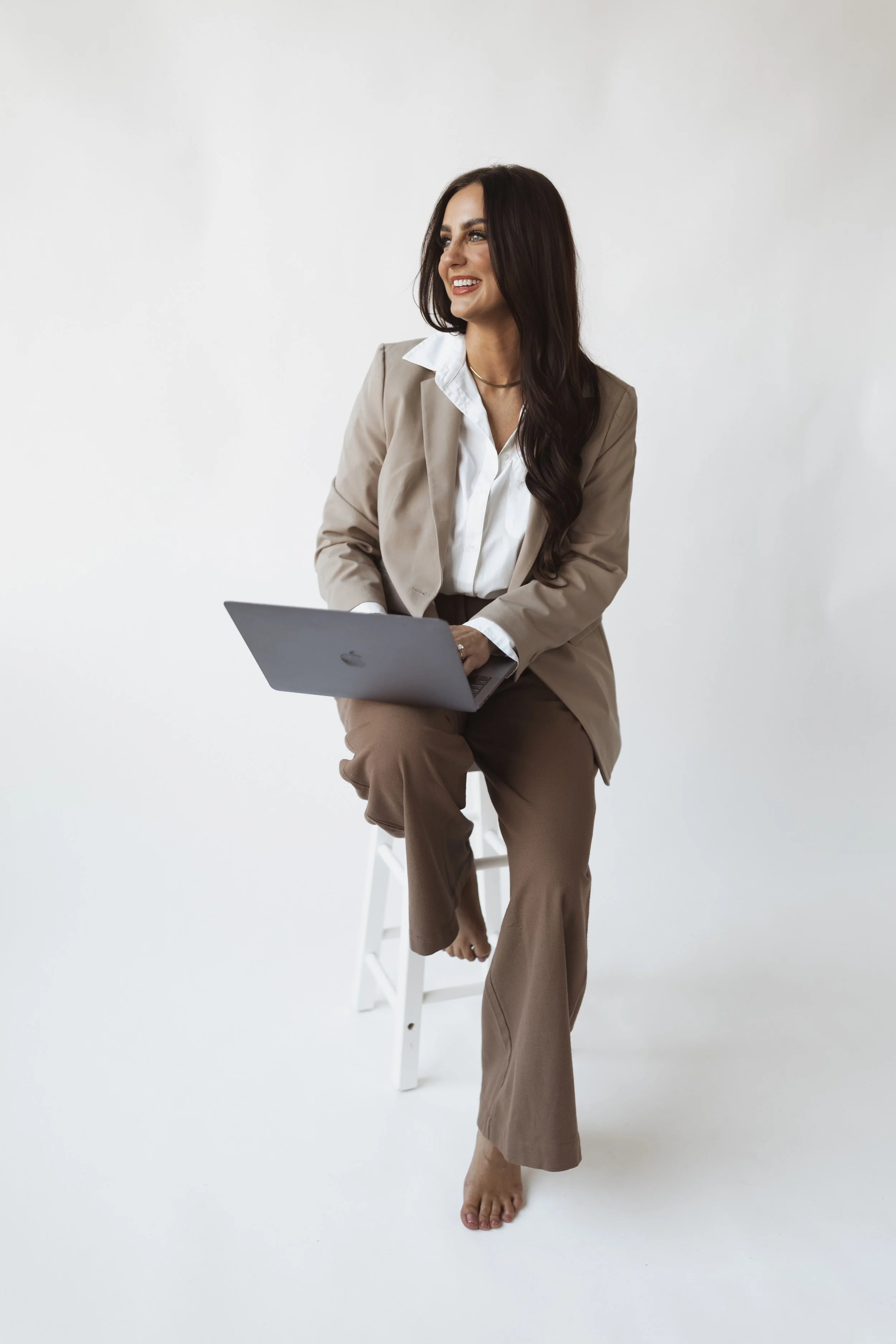 A woman in beige suit and white shirt sitting on a stool, holding a laptop, barefoot, smiling against a plain white background.