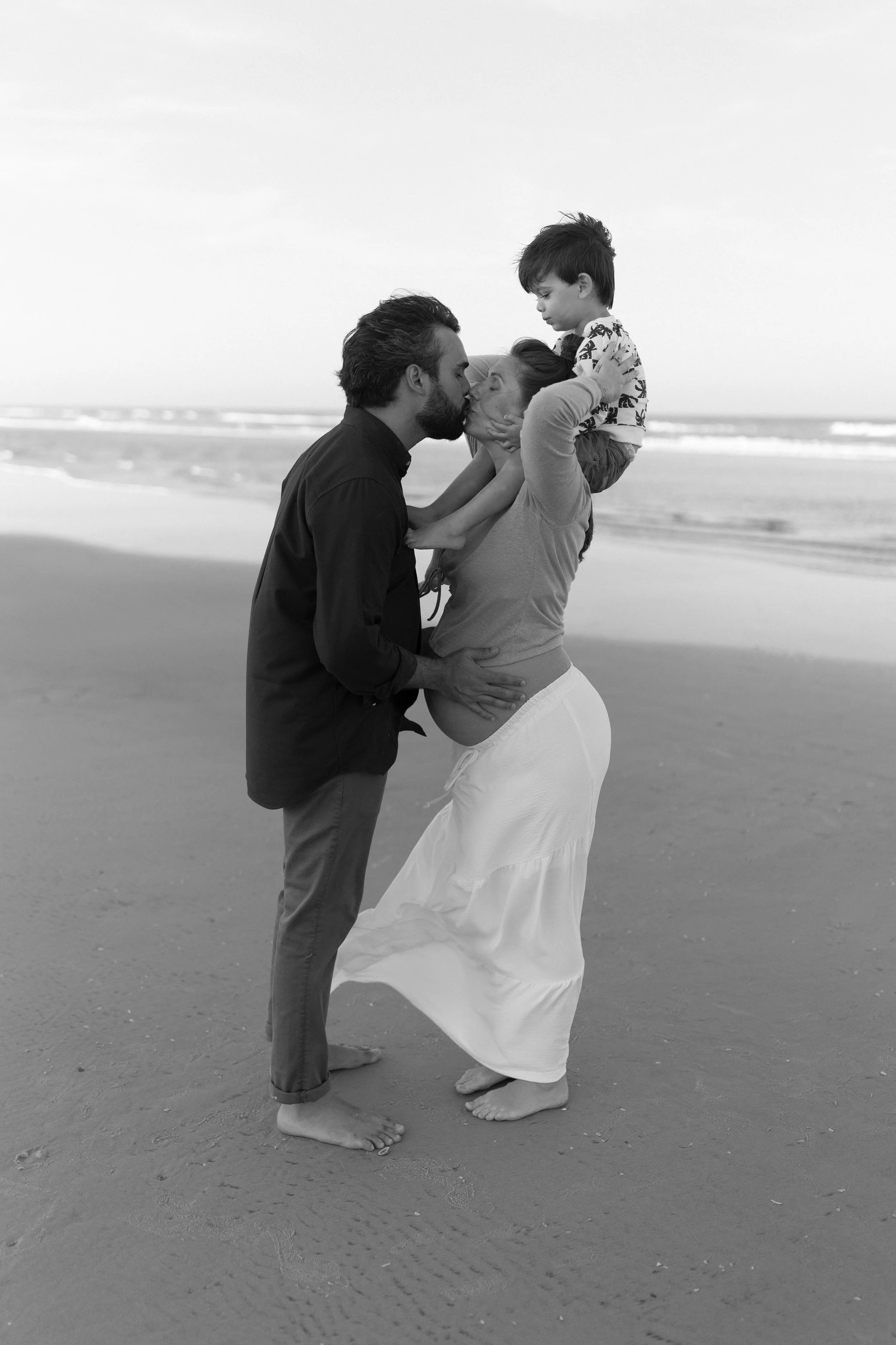 A black-and-white photo of a family on a beach. A woman with long hair and a long skirt is holding a young boy on her shoulders, and they are about to kiss a man with a beard who is also holding her around the waist.
