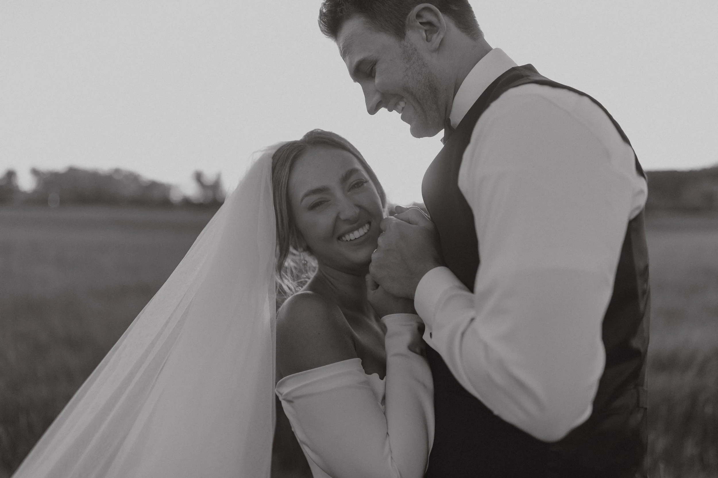 A smiling bride and groom are holding hands and looking into each other's eyes during sunset. The bride is wearing a wedding dress and veil, and the groom is in a tuxedo with a vest. They are outdoors in a field.