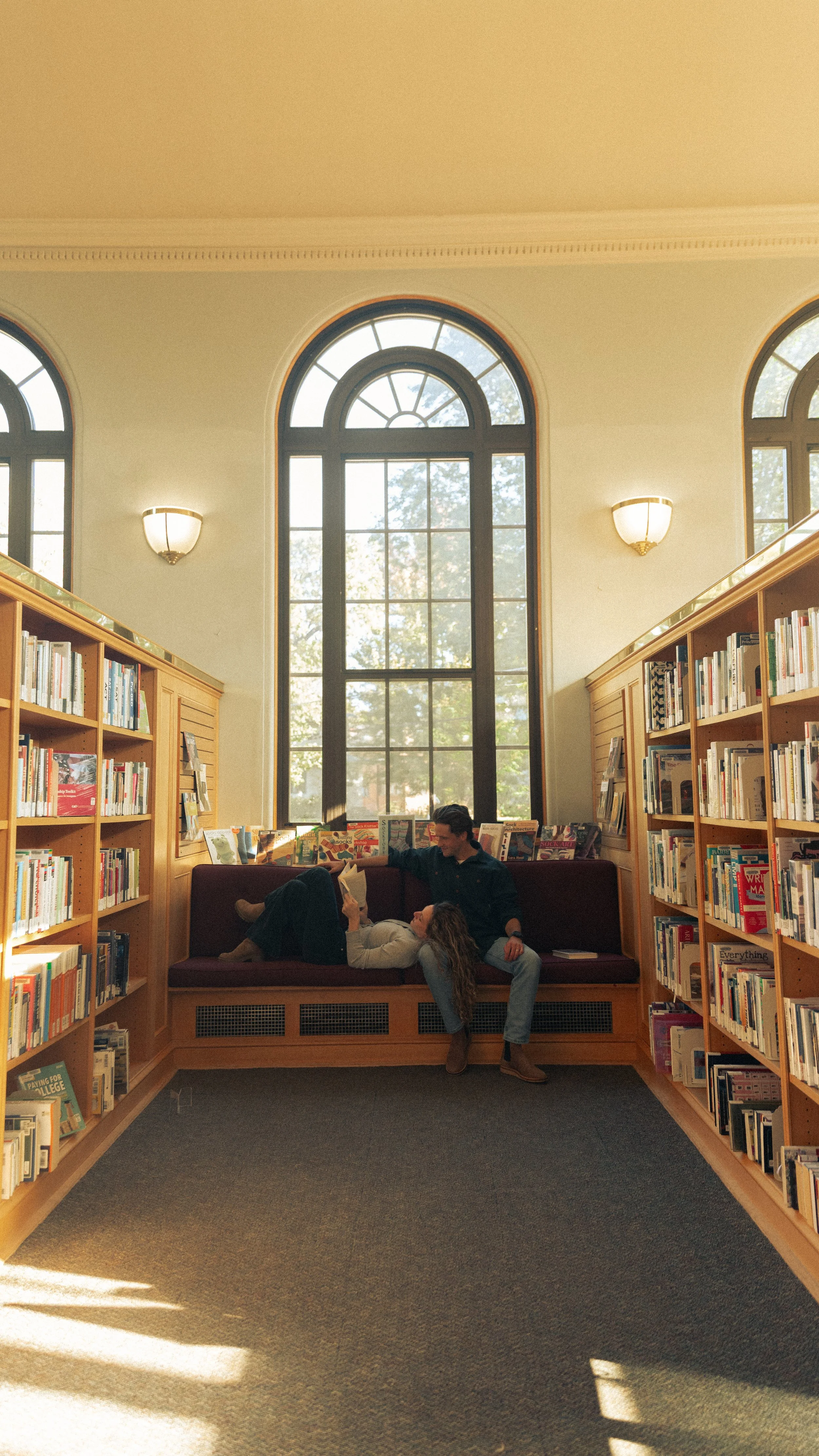Two people relaxing and reading in a cozy corner of a public library, surrounded by bookshelves and large arched windows.