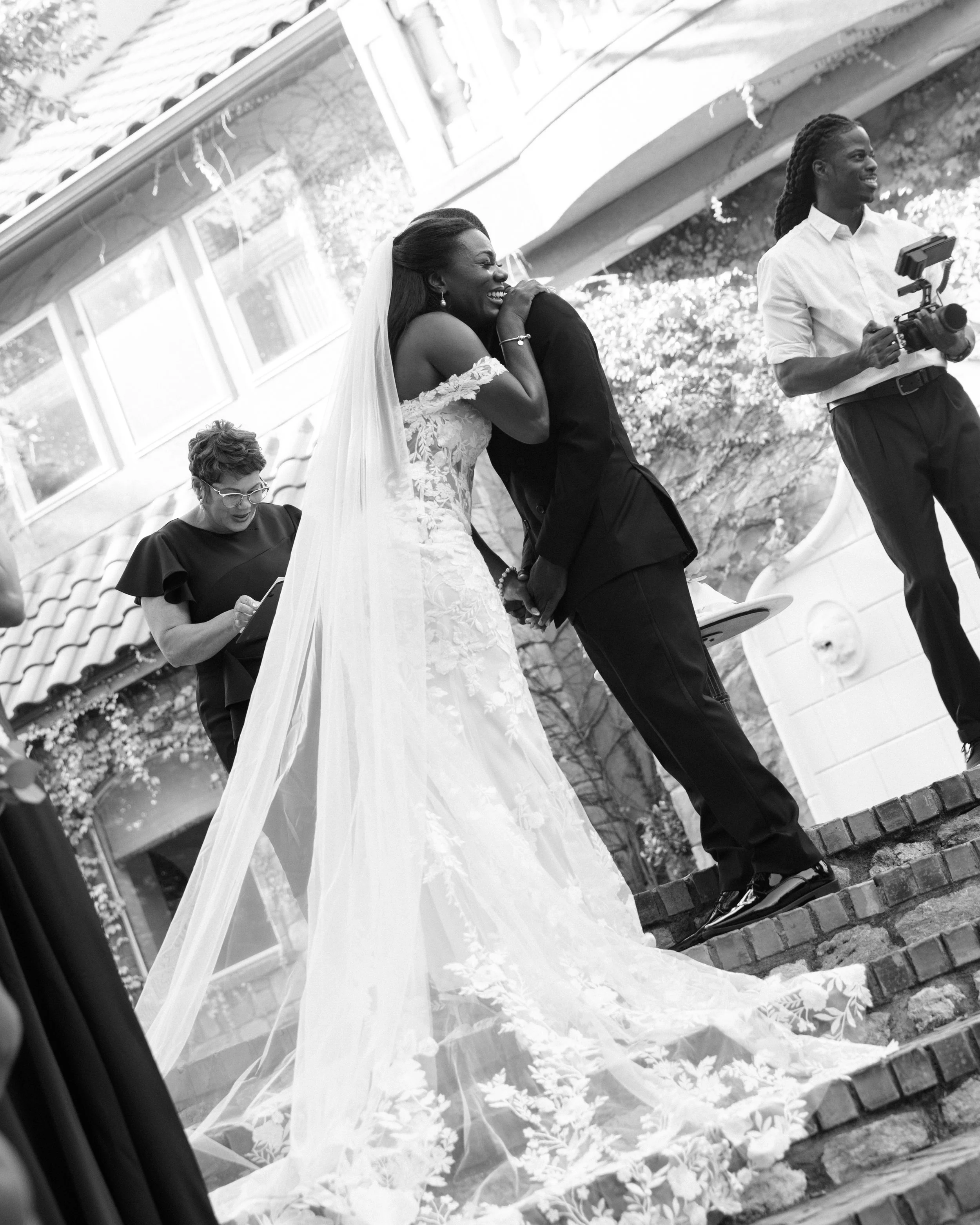 Black and white photograph of a bride and groom sharing a hug outdoors during their wedding ceremony. The bride is wearing a lace wedding gown with a long veil, and the groom is in a dark suit. A photographer is taking pictures nearby, and a woman wi