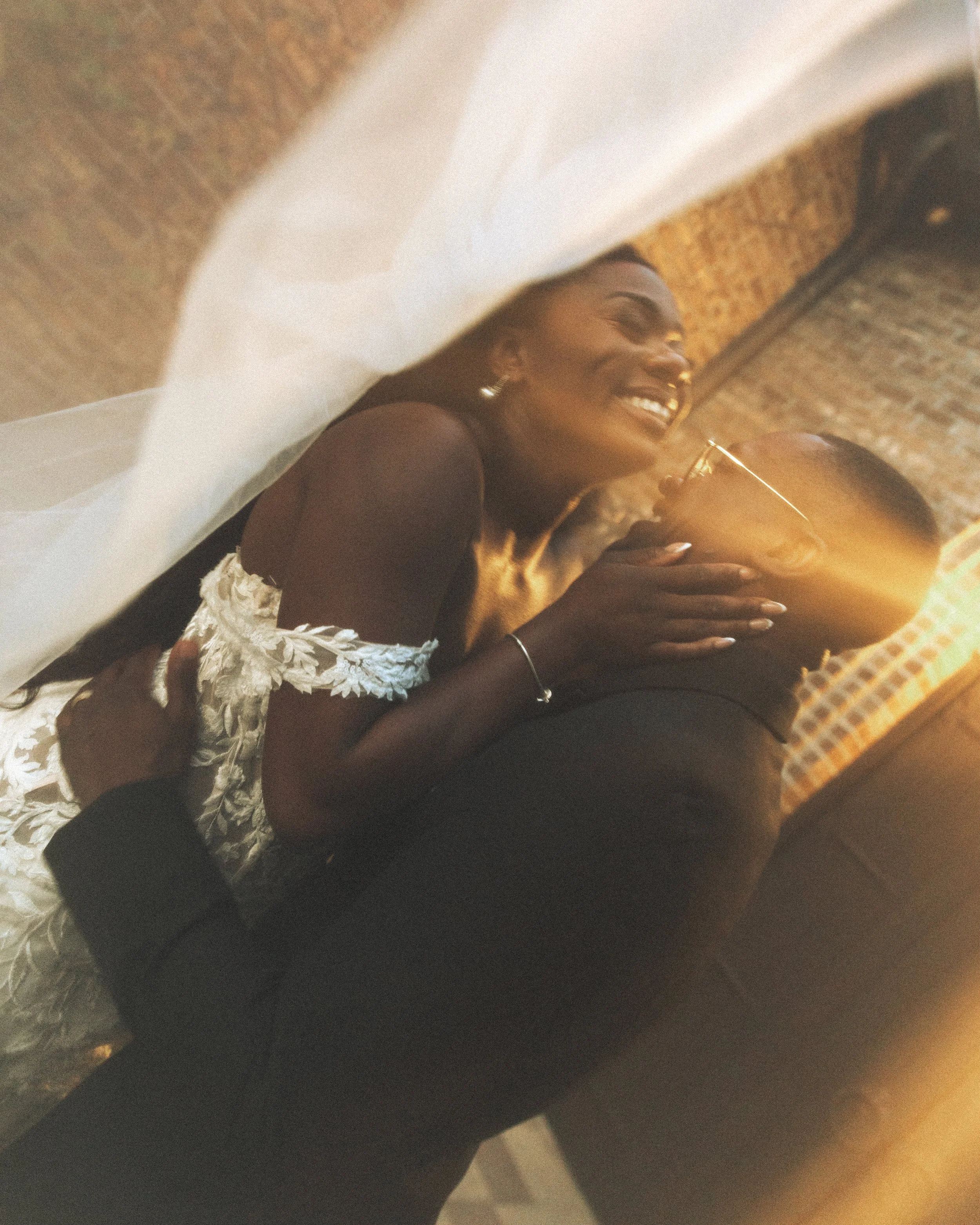 A woman with long dark hair, dressed in a white lace dress, is lying upside down on a man's shoulders and smiling, with sunlight shining in the background.