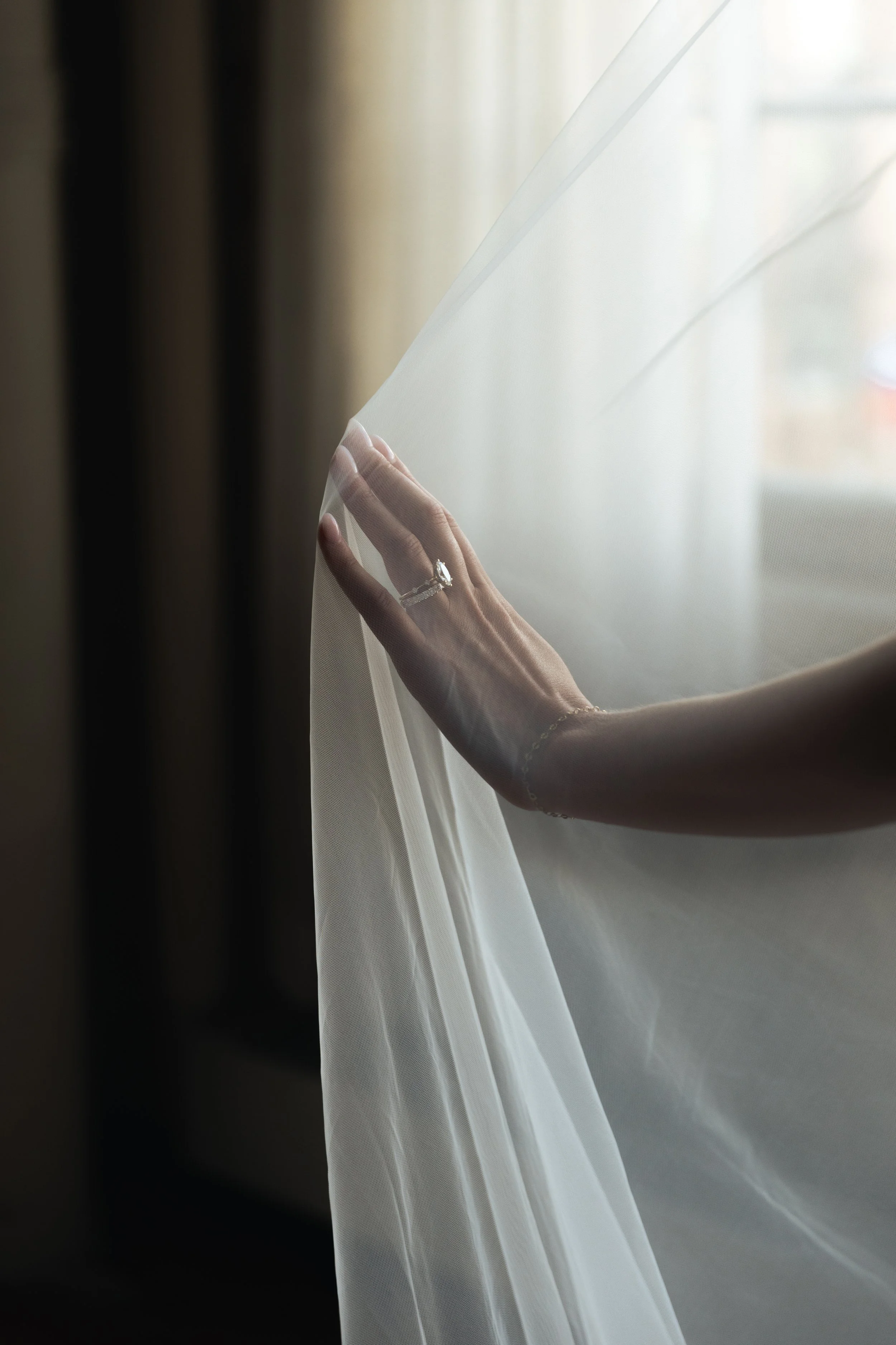 A bride's hand touching a sheer curtain, showing an engagement ring with a large center stone and a wedding band.