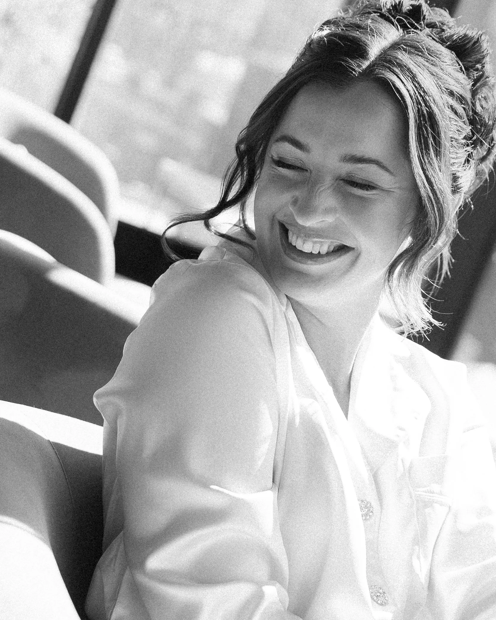 A woman smiling and looking to her left with short wavy hair, wearing a blouse, sitting in a well-lit room with chairs in the background.