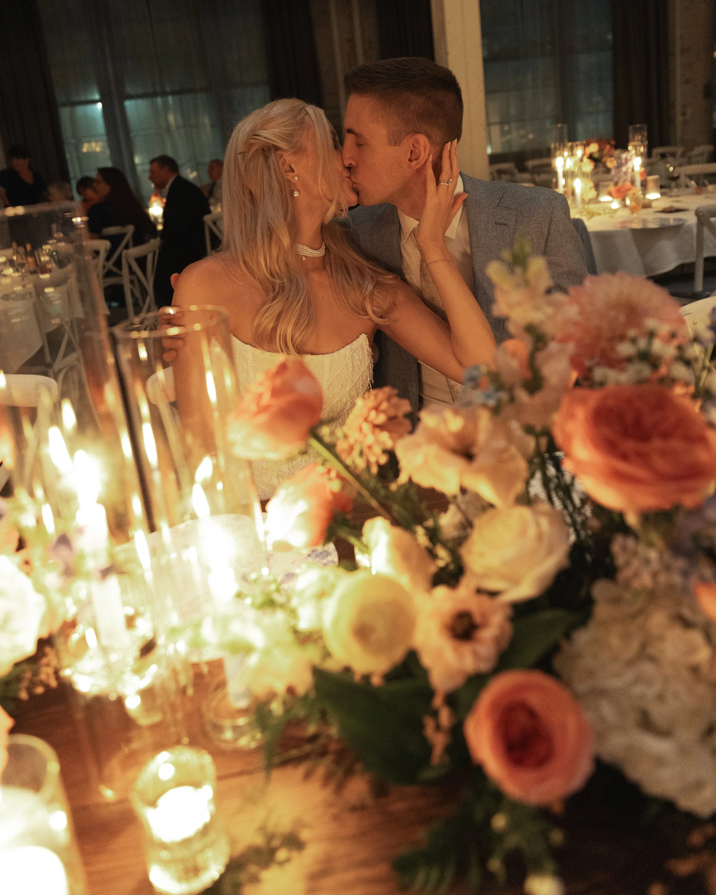 A couple kissing at a wedding reception with floral arrangements and candlelit tables in the background.