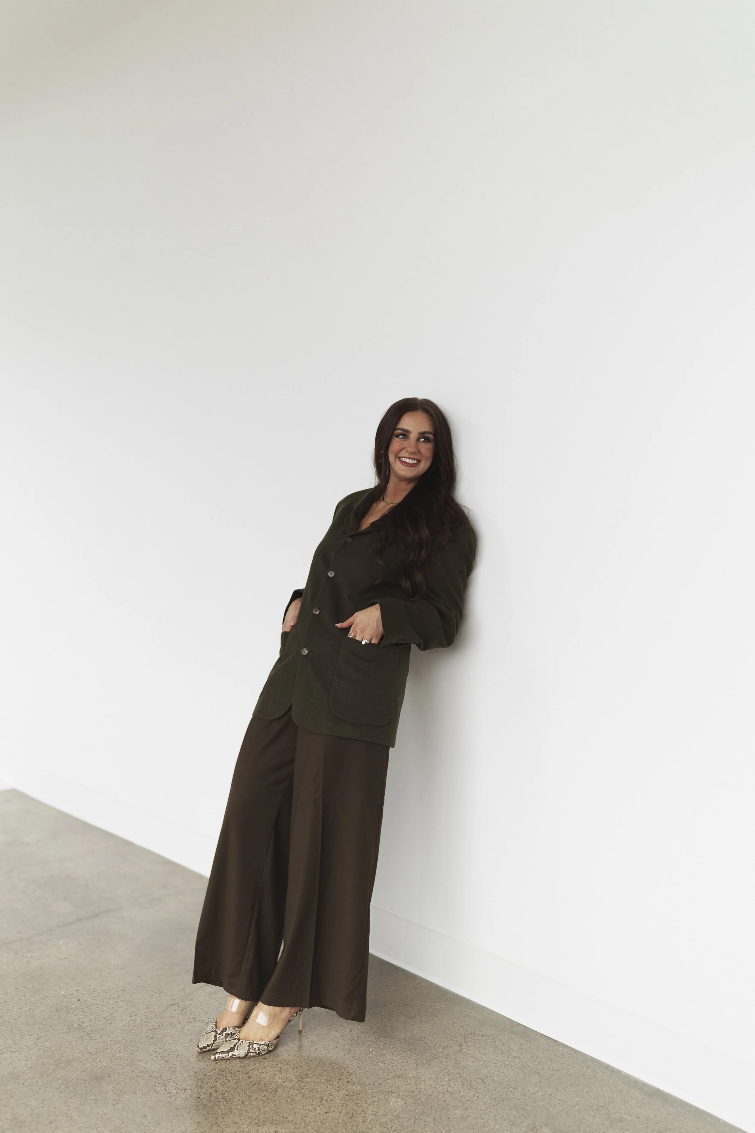 Woman with long dark hair smiling, standing against a white wall, wearing a dark green blazer, matching wide-leg pants, and patterned high heels.