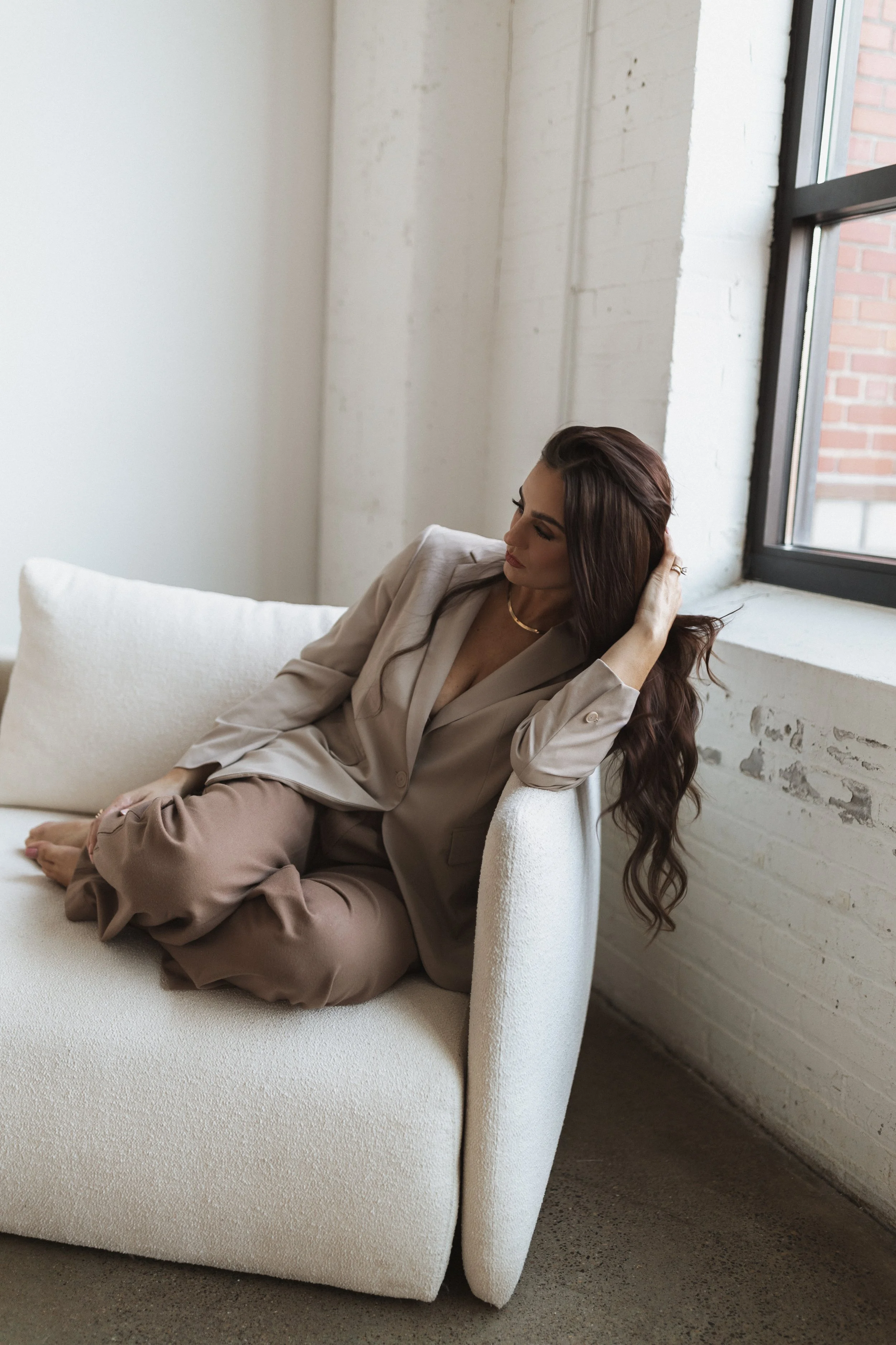 A woman in beige pajamas relaxing on a white couch near a window in a modern, minimalist room.