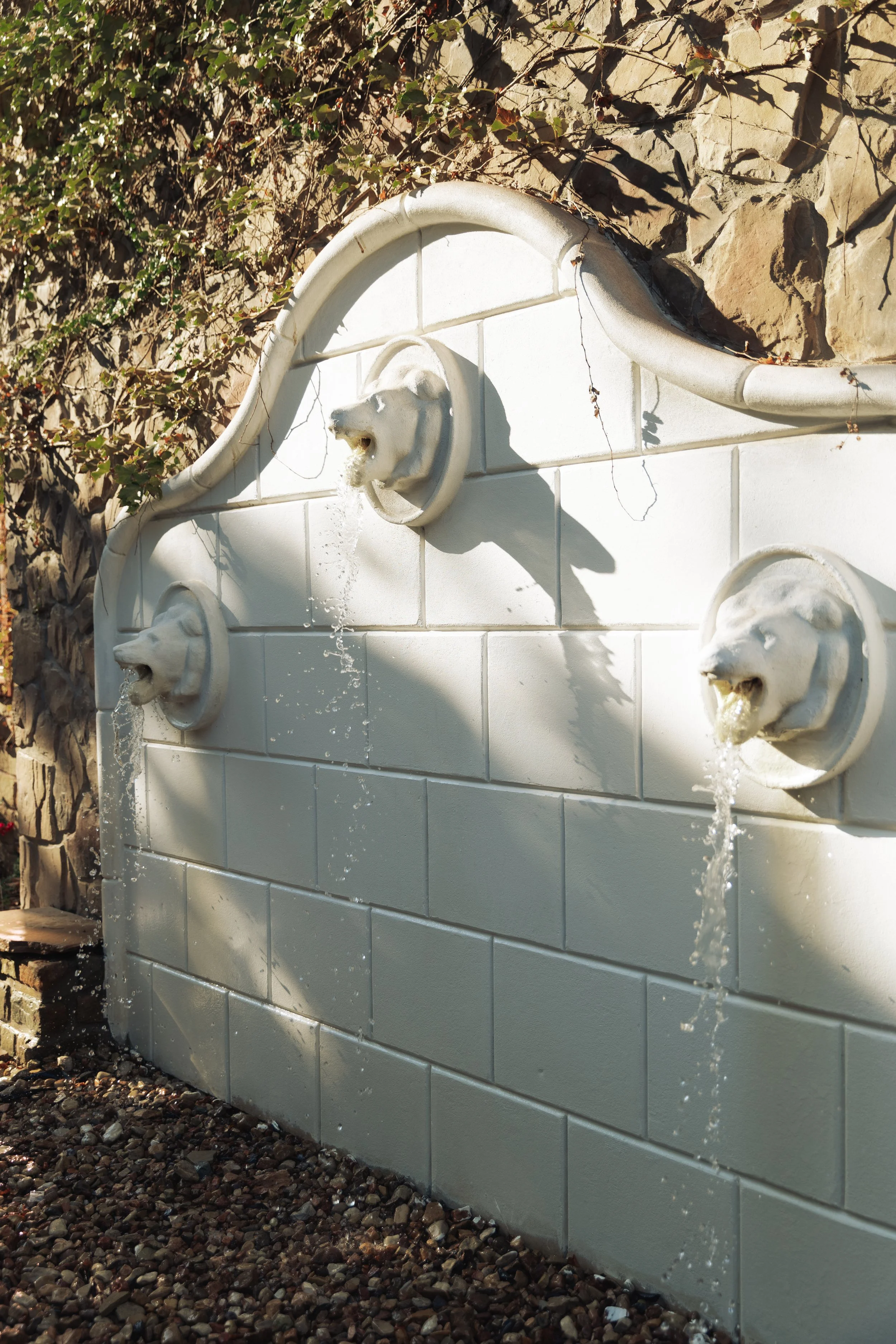 Stone wall with three lion head water fountains, each with water flowing from the mouth, against a backdrop of ivy and rocks.