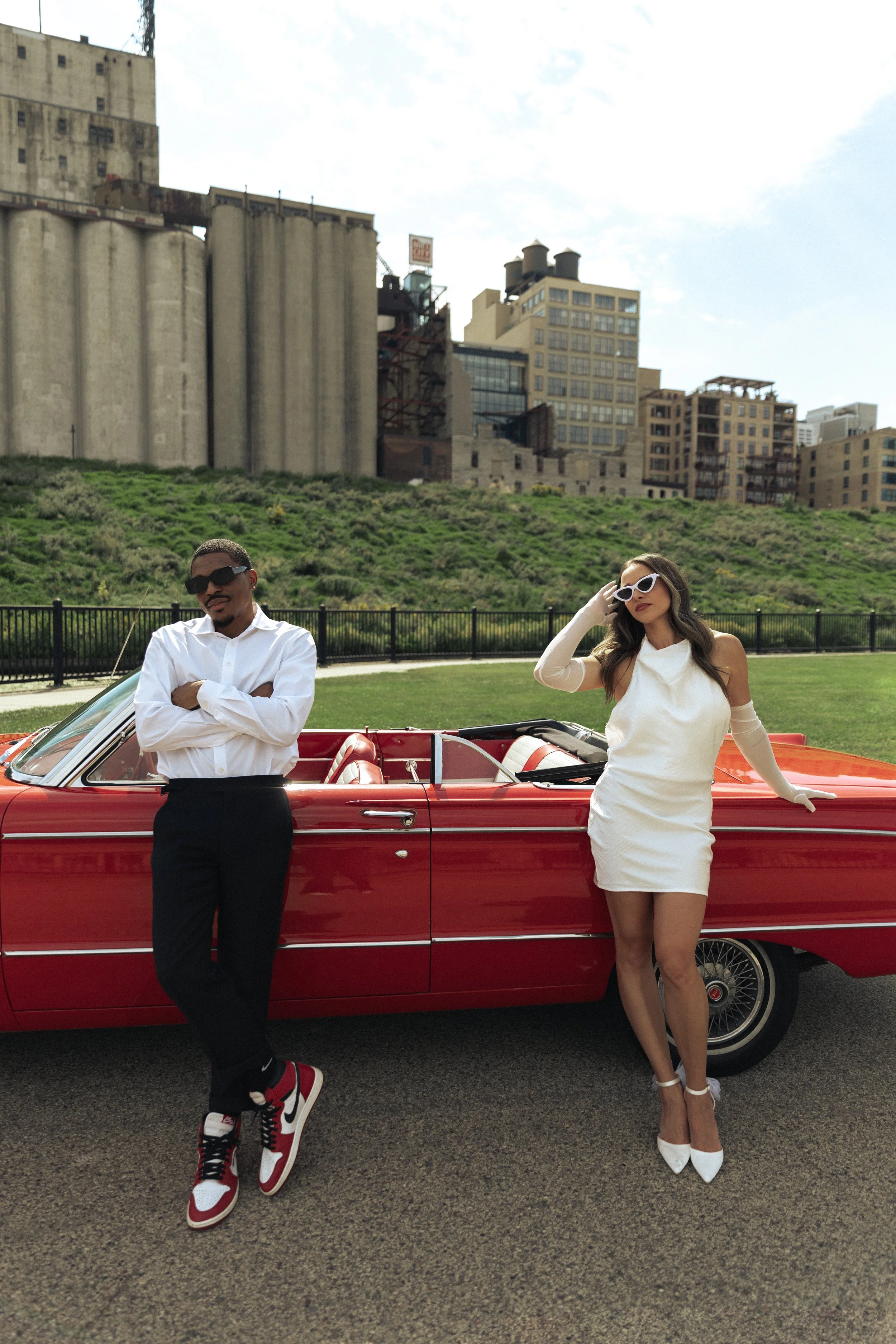 A man in a white shirt and black pants leaning against a red vintage convertible car, and a woman in a white dress and high heels leaning on the same car, both wearing sunglasses, in an outdoor urban park with city buildings in the background.