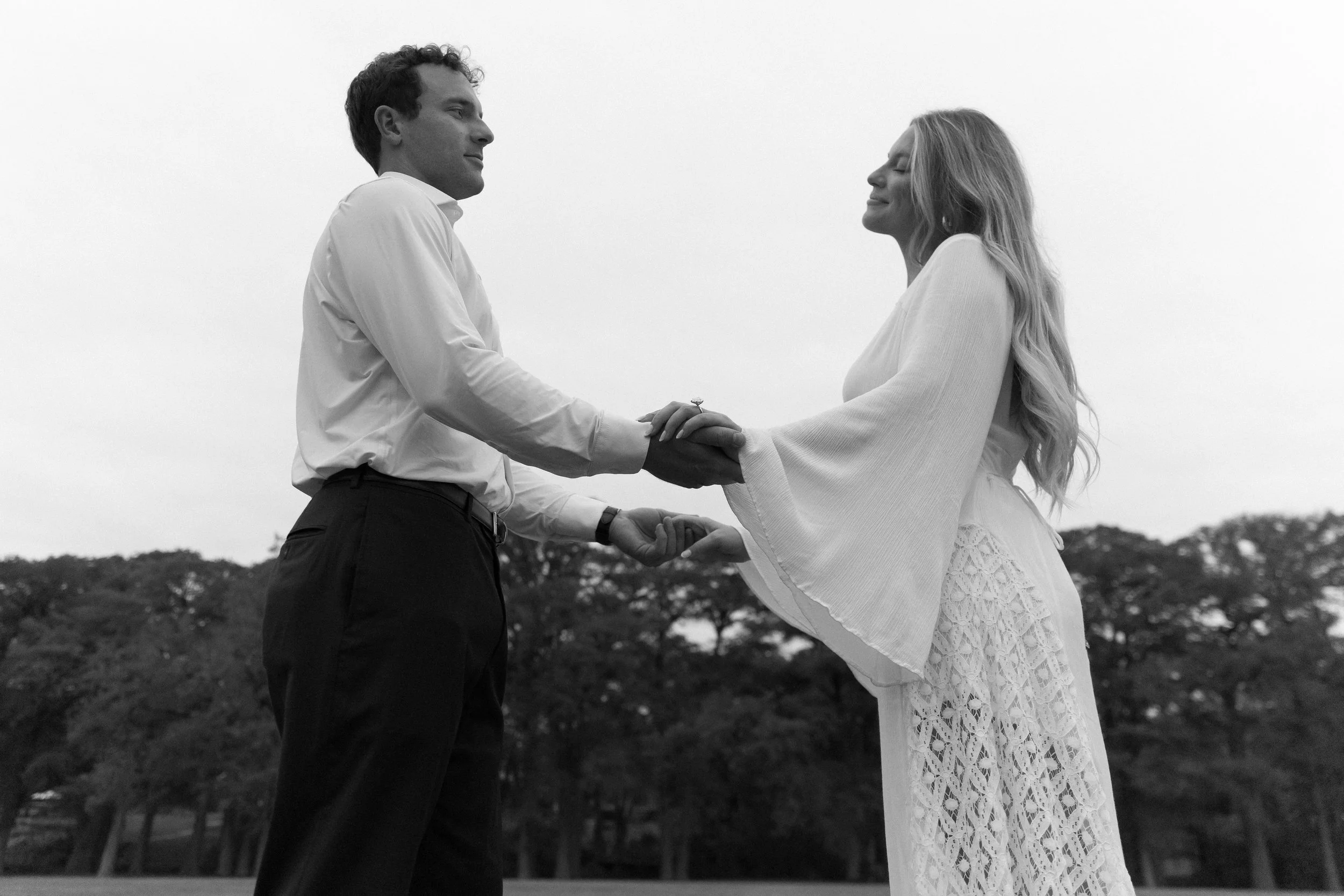 A man and woman holding hands in a romantic outdoor setting, with the man wearing a white shirt and dark pants, and the woman dressed in light-colored lace clothing, both smiling.