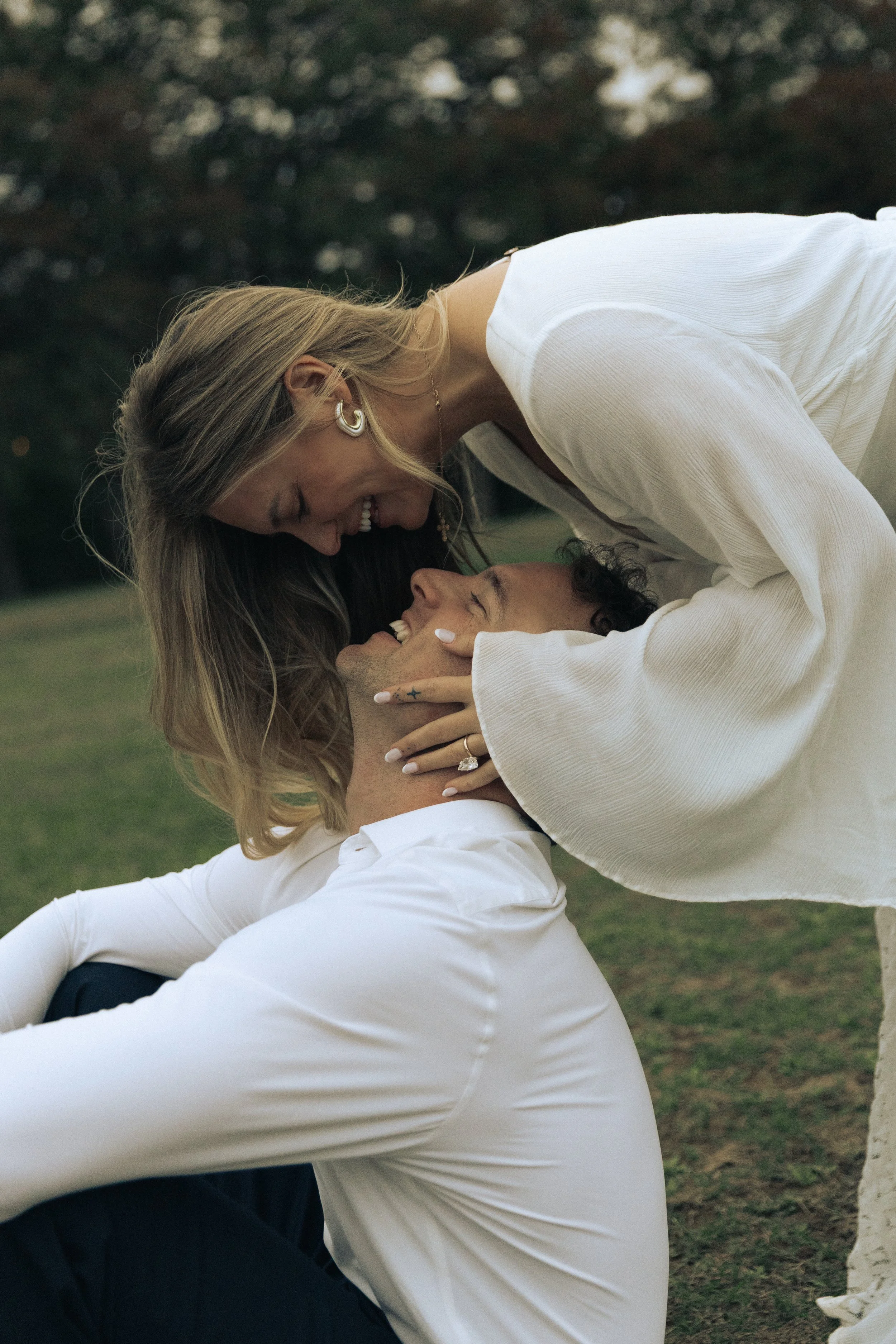 A couple outdoors, smiling and holding each other closely, with the woman leaning over the man, in a park setting during daytime.