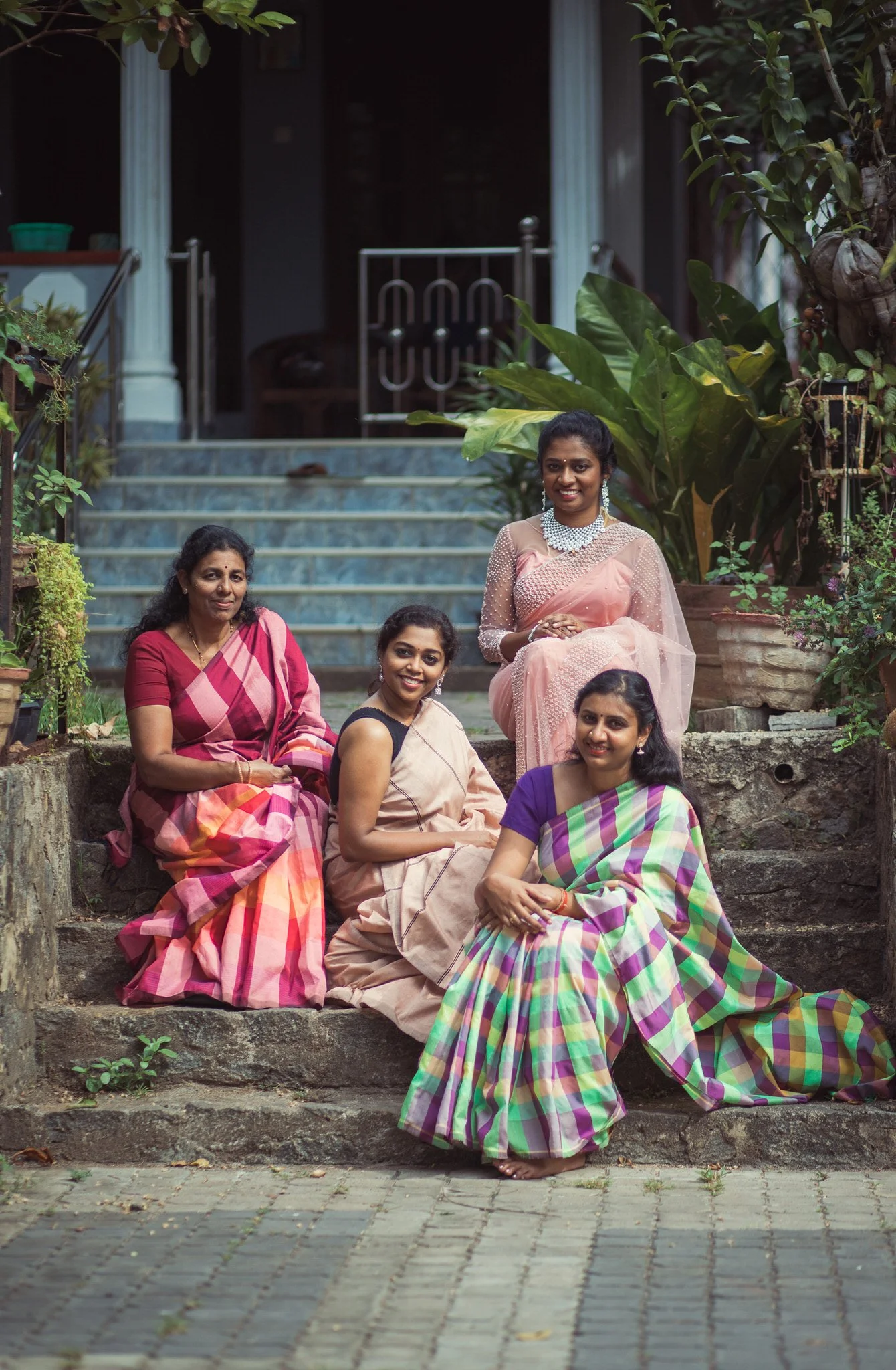 Five women sitting on stone steps outside a house, dressed in traditional sarees with colorful patterns, surrounded by potted plants and greenery.