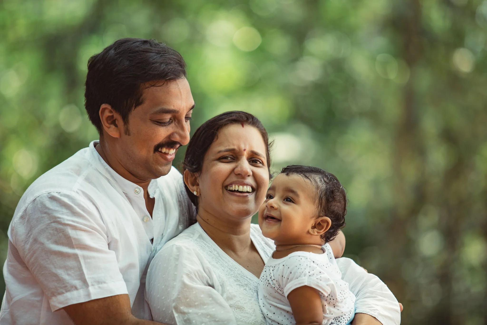 A smiling South Asian family of three enjoying time outdoors in a green, blurred background. The father, mother, and toddler are all smiling and looking happy.