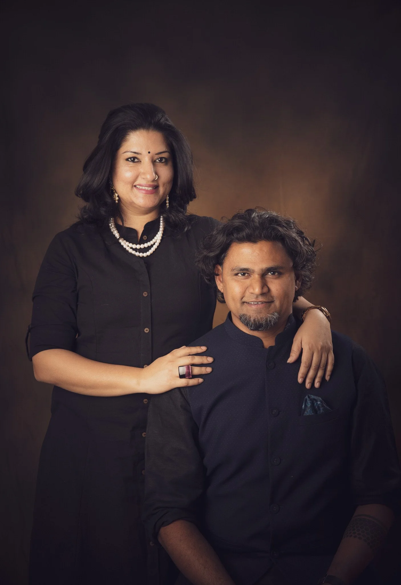 A woman with black hair, wearing a black top and pearl jewelry, standing behind a man with curly hair, wearing a dark shirt, both smiling in a studio portrait.