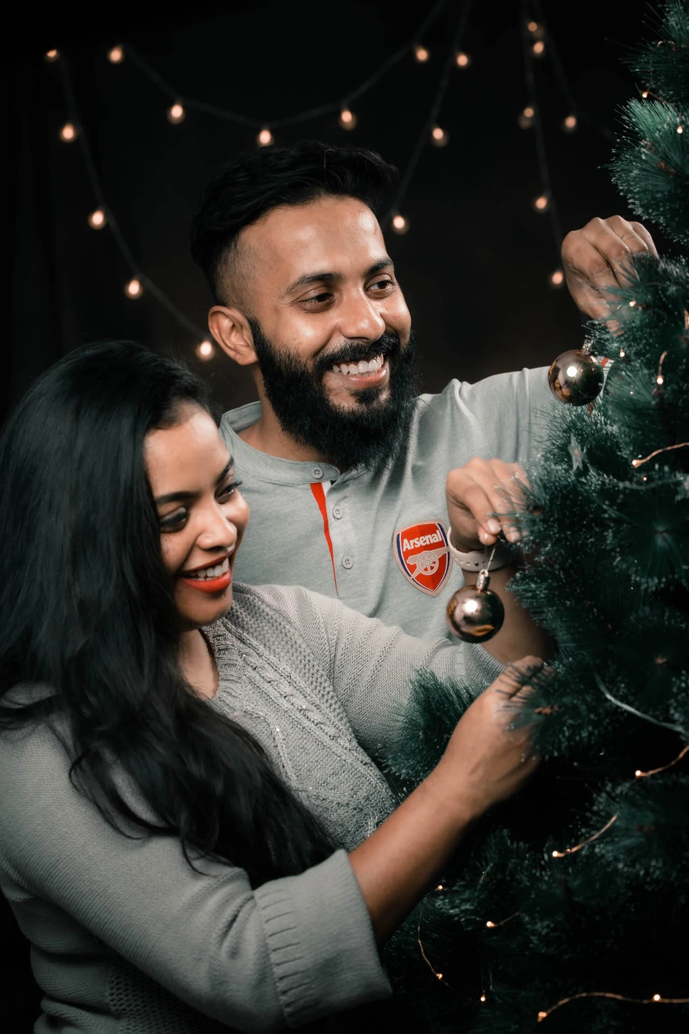 A man and woman decorating a Christmas tree together, smiling, with a dark background and string lights.