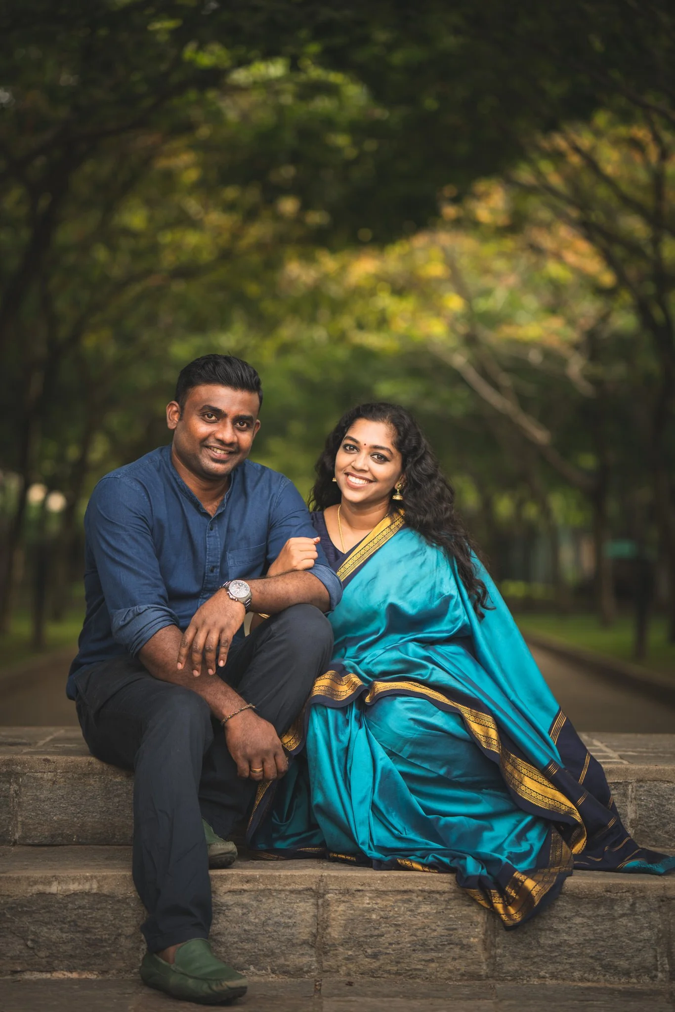 A man and woman sitting on stairs outdoors, smiling, with trees in the background. The woman is wearing a blue saree with gold accents, and the man is wearing a navy blue shirt and black pants.