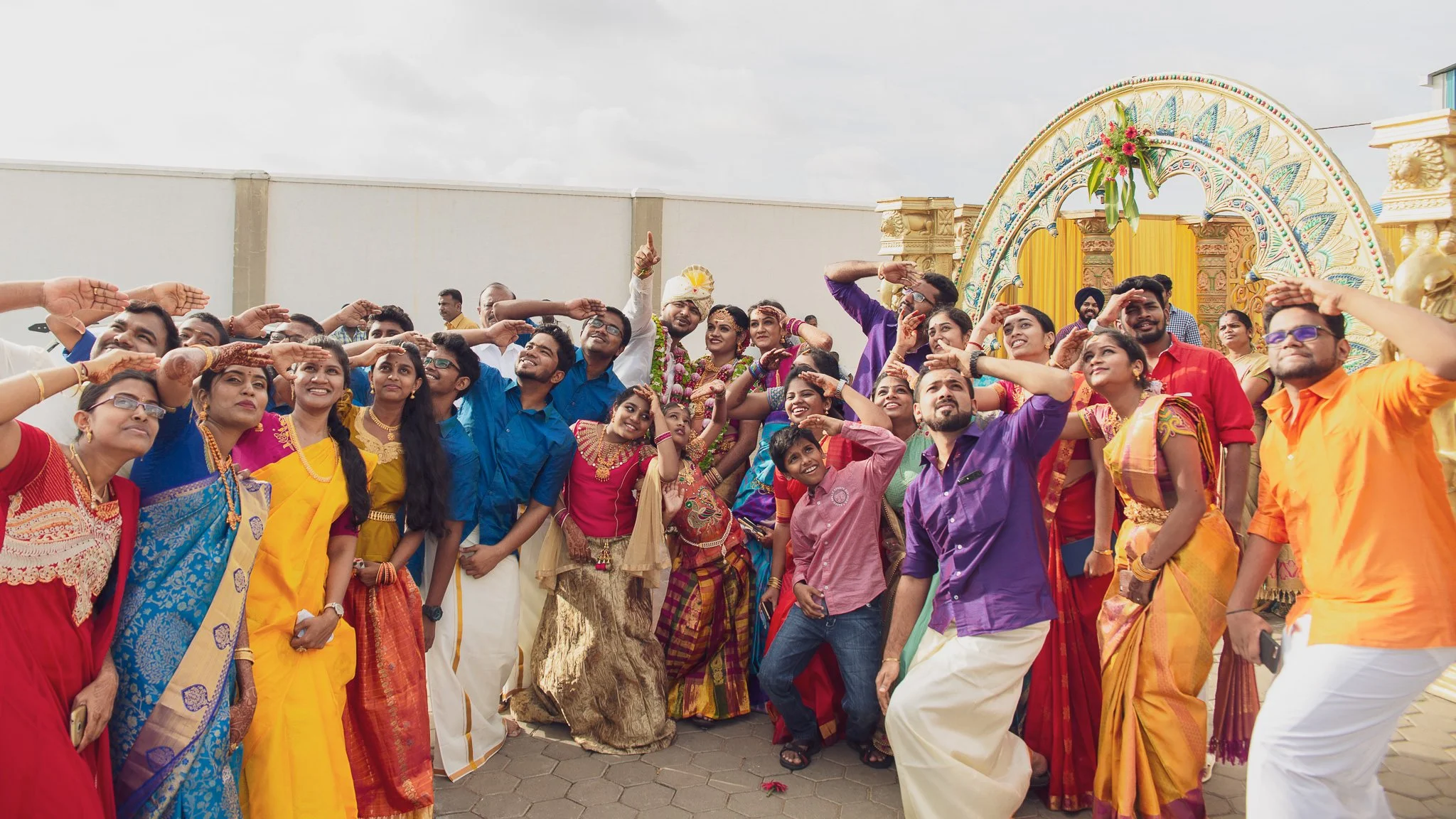 A group of people dressed in colorful traditional Indian clothing saluting in front of a decorated arch at a celebration or wedding.