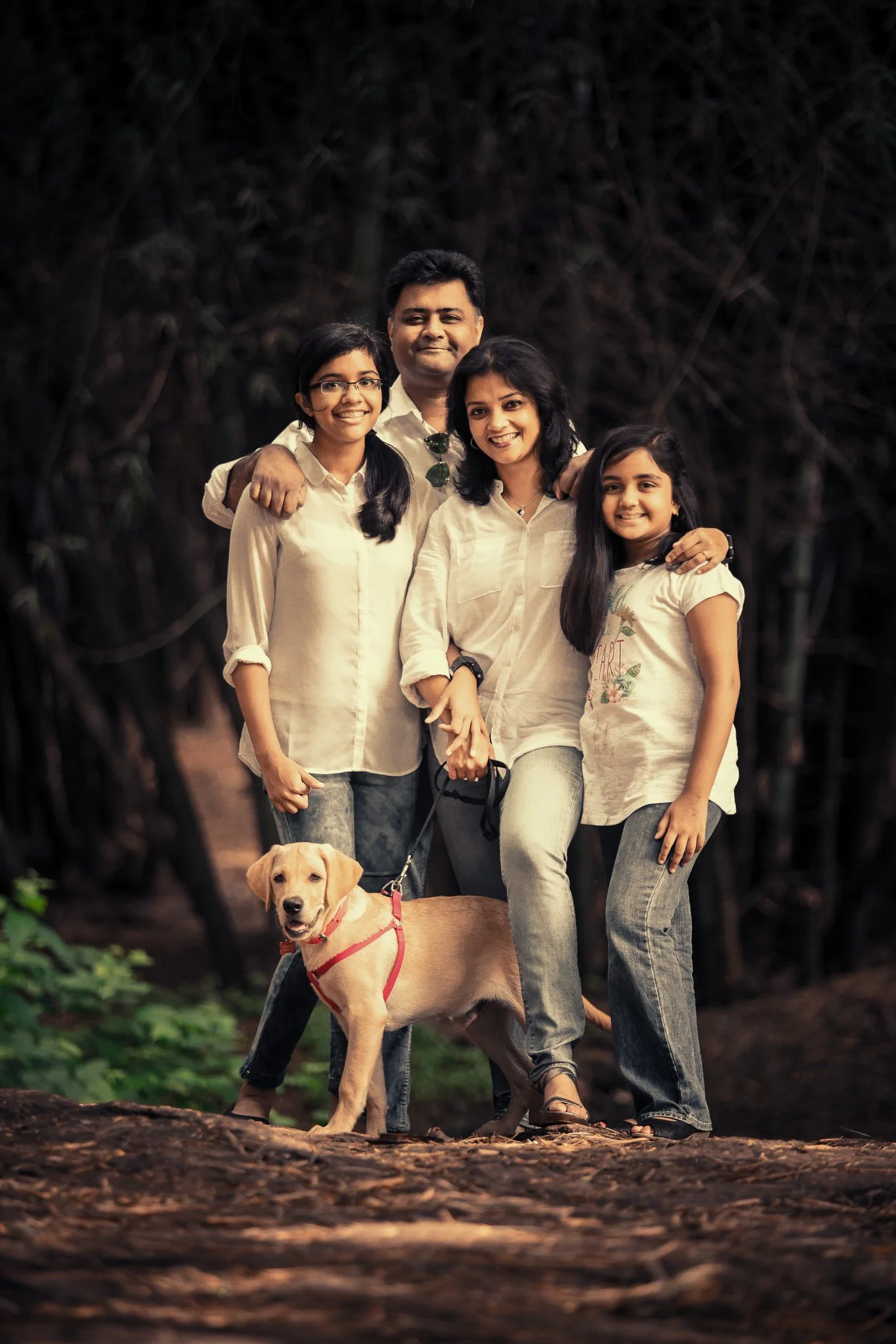 A family of five, including a dog, taking a group photo outdoors in a wooded area during daytime. The family consists of a man, a woman, two girls, and a Labrador retriever with a red harness. They are all dressed casually and smiling.