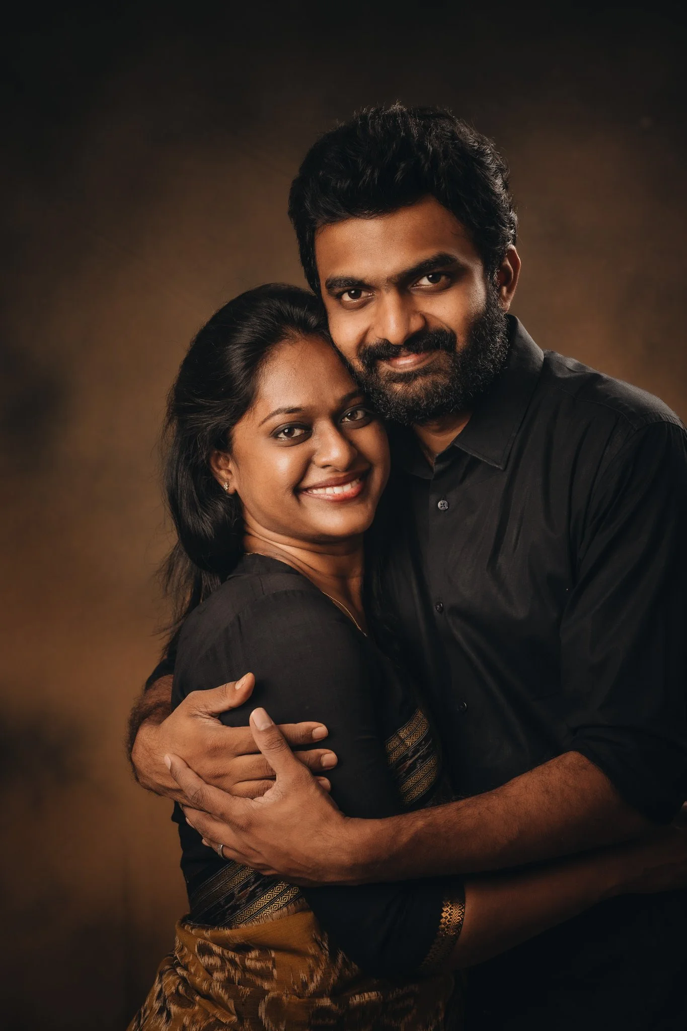 A smiling woman and man embracing, looking at the camera against a brown studio background.
