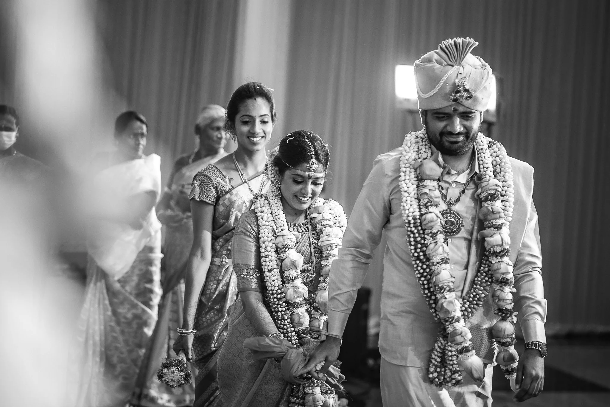 A black and white photo of a traditional Hindu wedding ceremony with the bride and groom holding hands, walking in front of a group of women dressed in sarees, inside a decorated hall.