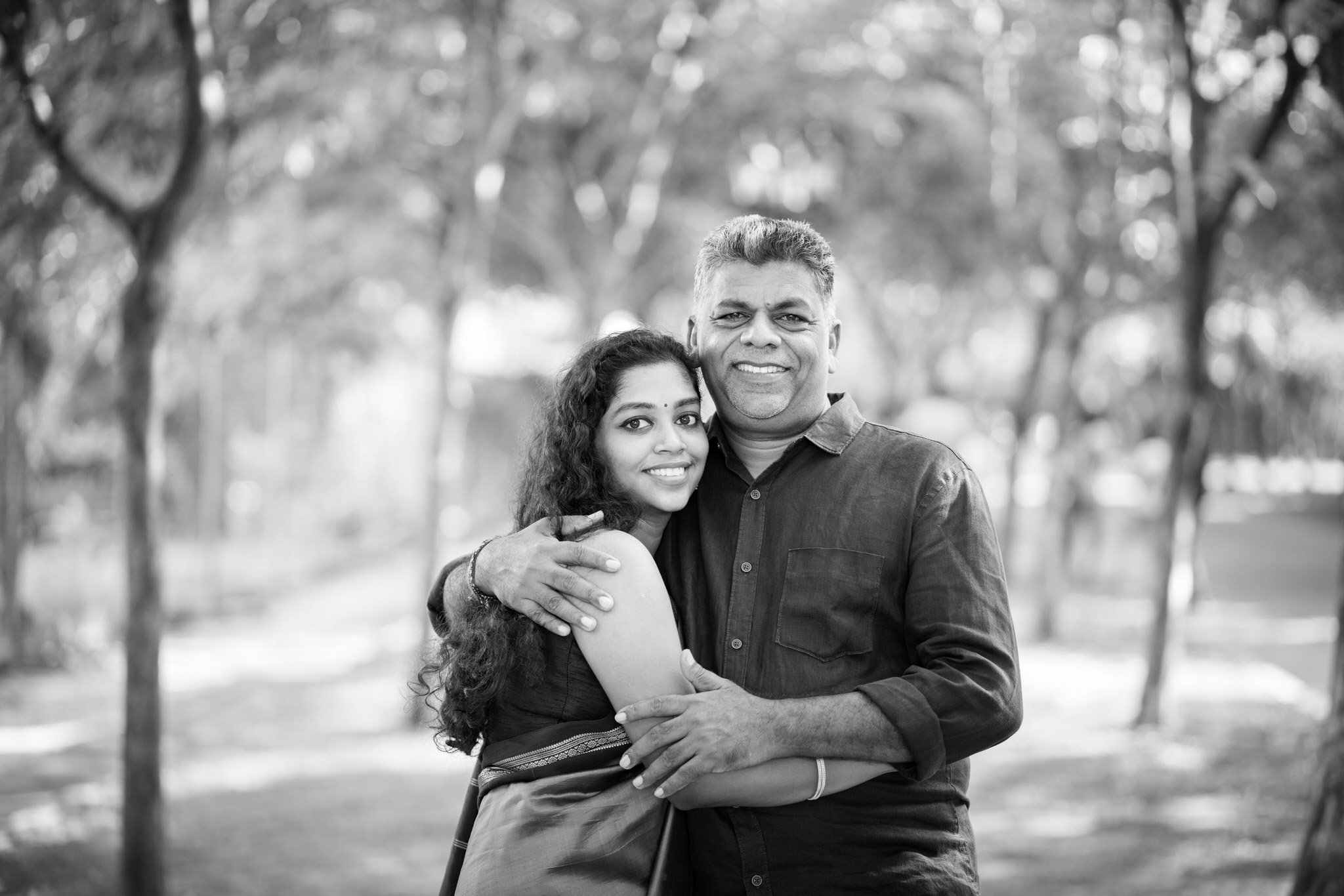 A black and white photo of a smiling couple standing outdoors with trees in the background, embracing each other.