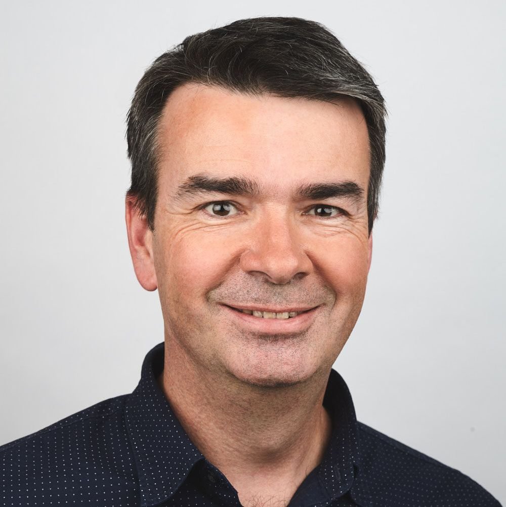 A professional headshot of a middle-aged man with dark hair, smiling, wearing a dark collared shirt, against a plain light background.