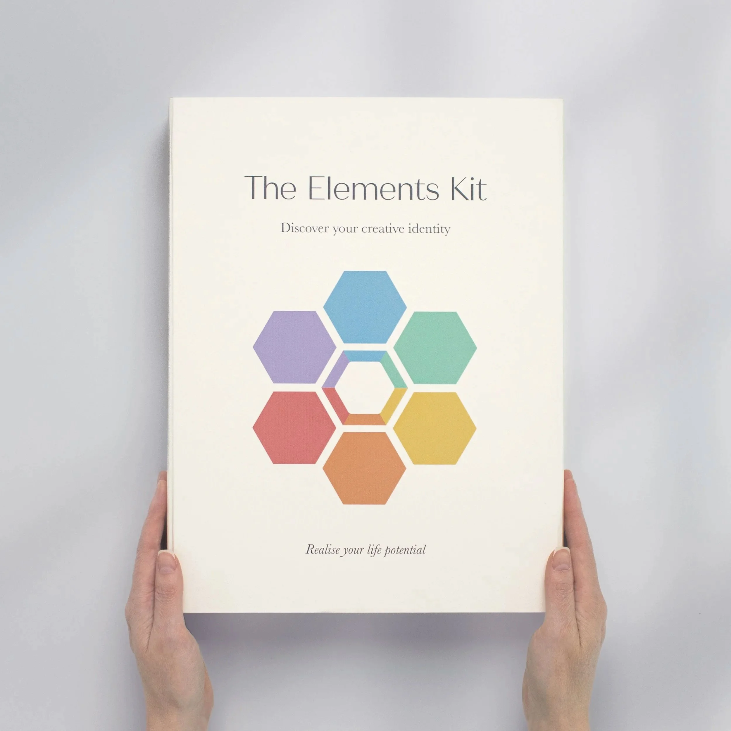 A person holding a white box labeled "The Elements Kit" with a colorful hexagon design and the text "Discover your creative identity" and "Realise your life potential."