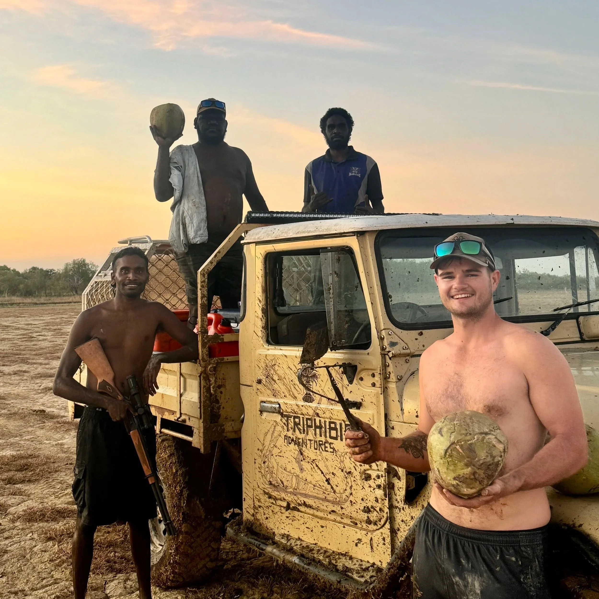 Four men standing around a 40 series landcruiser in a muddy terrain during sunset, holding coconuts and a rifle, with a cloudy sky in the background.