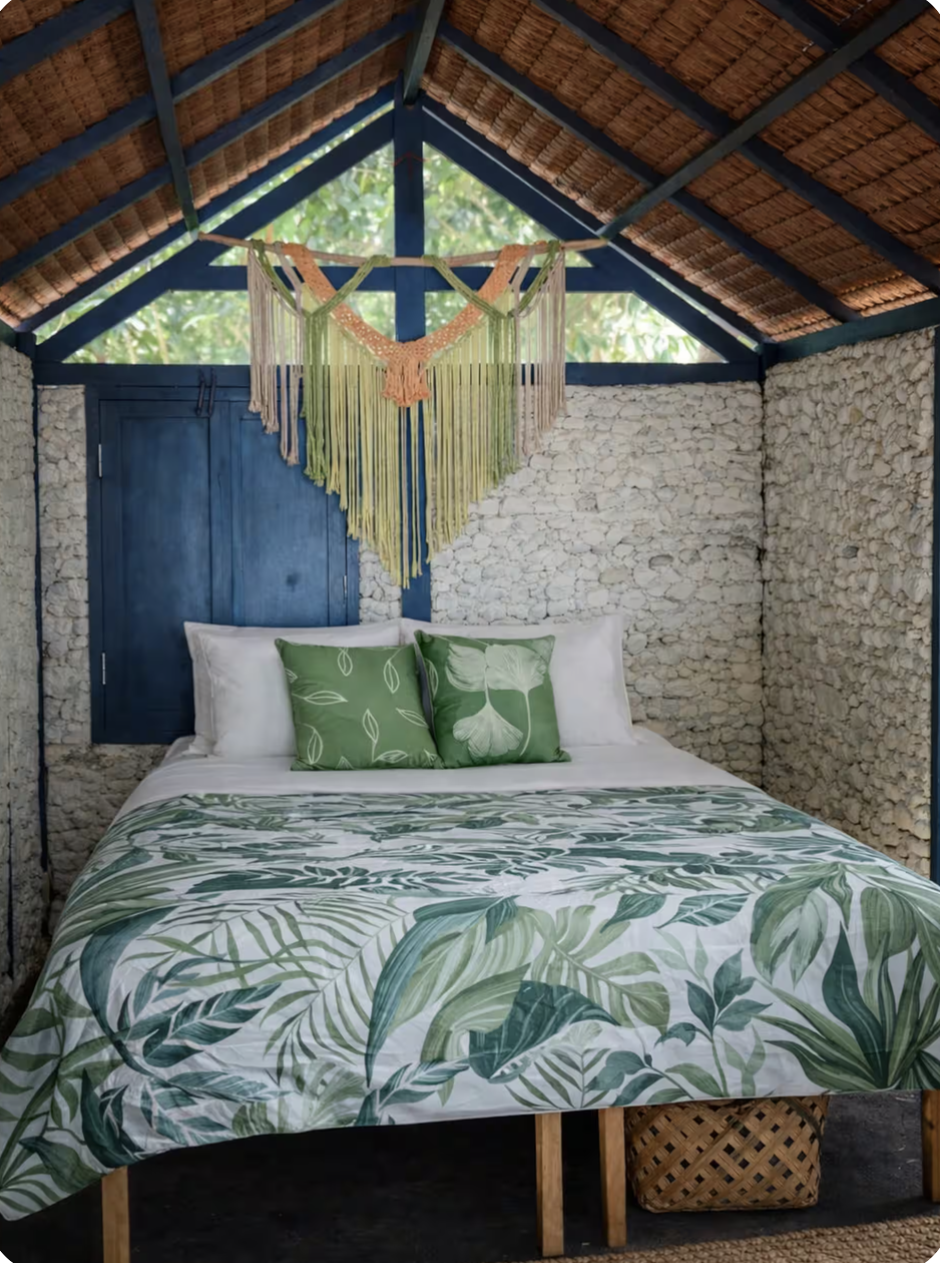 A rustic bedroom with stone walls and a high thatched roof. A bed with tropical leaf-patterned bedding and two green pillows is centered in the room. Above the bed, a colorful macrame wall hanging is displayed, with a blue door visible behind the pil
