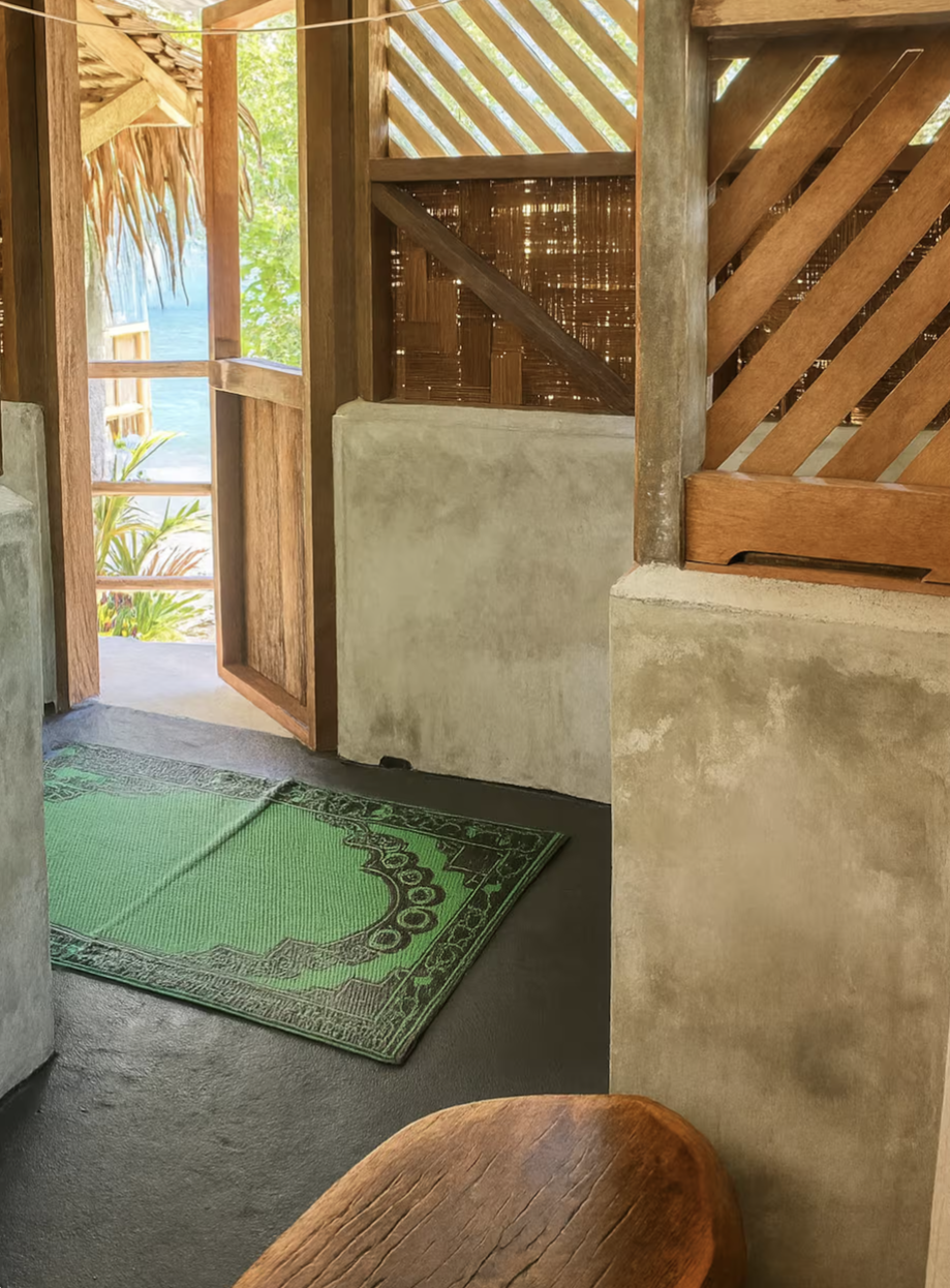 Interior of a rustic outdoor hut with wooden walls, a concrete half-wall, a green rug, and an open wooden door leading outside to tropical plants and water.