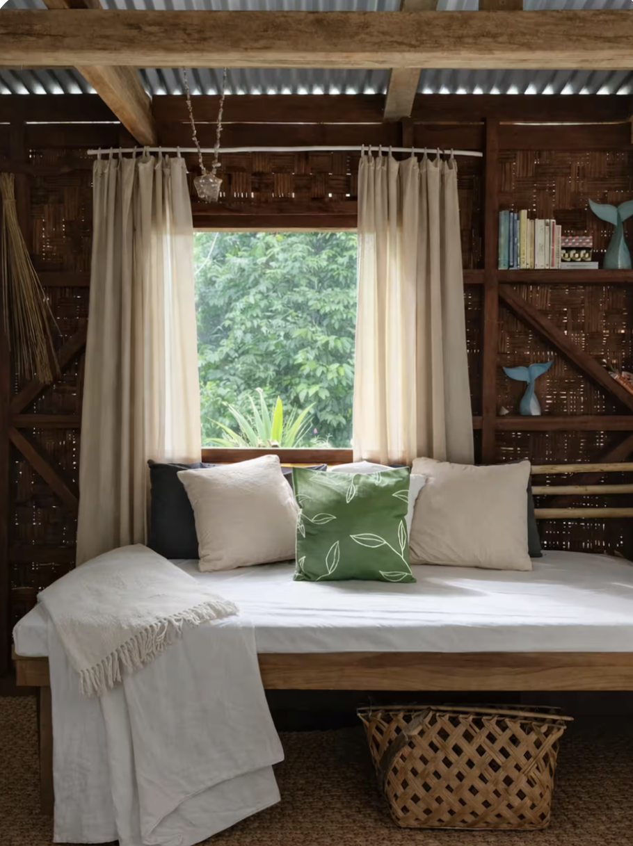 A cozy bedroom with a wooden bed frame, white bedding, and several pillows, including a green pillow with leaf patterns. Behind the bed, there's a window with cream curtains showing green foliage outside. To the right, wooden shelves hold books and d